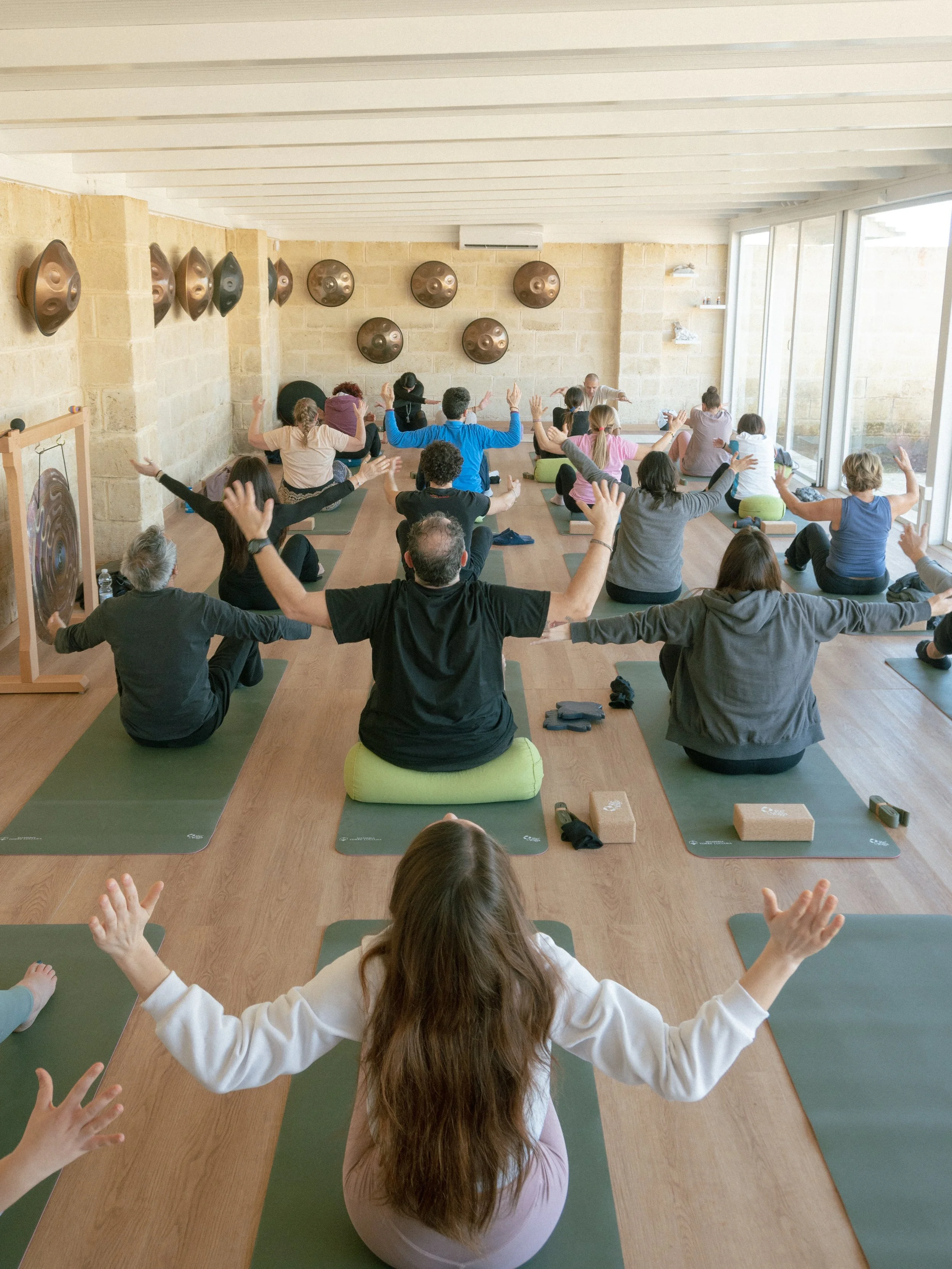 People participating in a yoga class in a spacious room with large windows and wooden floors, seated on mats with arms raised.
