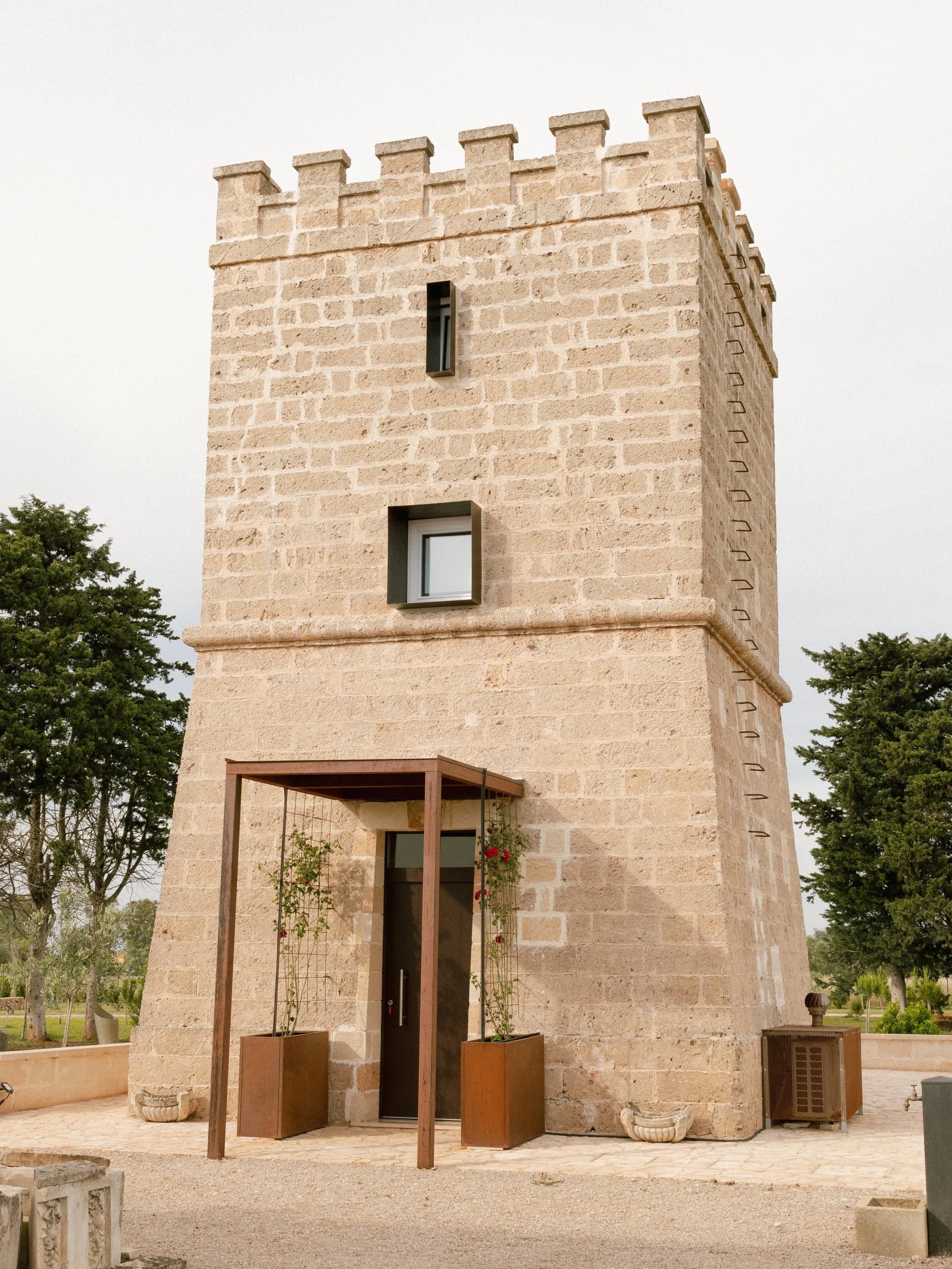 A tall stone tower with a crenellated top, small rectangular windows, and a modern entrance with potted plants on either side.