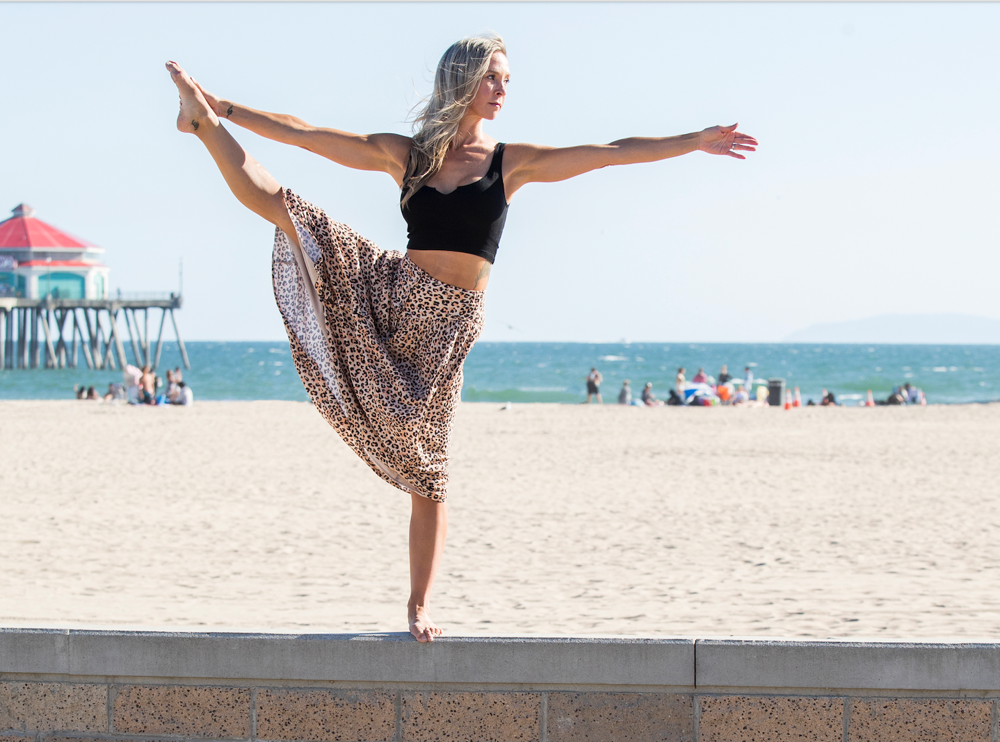 Woman practicing yoga on a ledge at the beach, balancing on one leg with arms outstretched, wearing a black crop top and leopard-print skirt, with the ocean and a pier in the background.