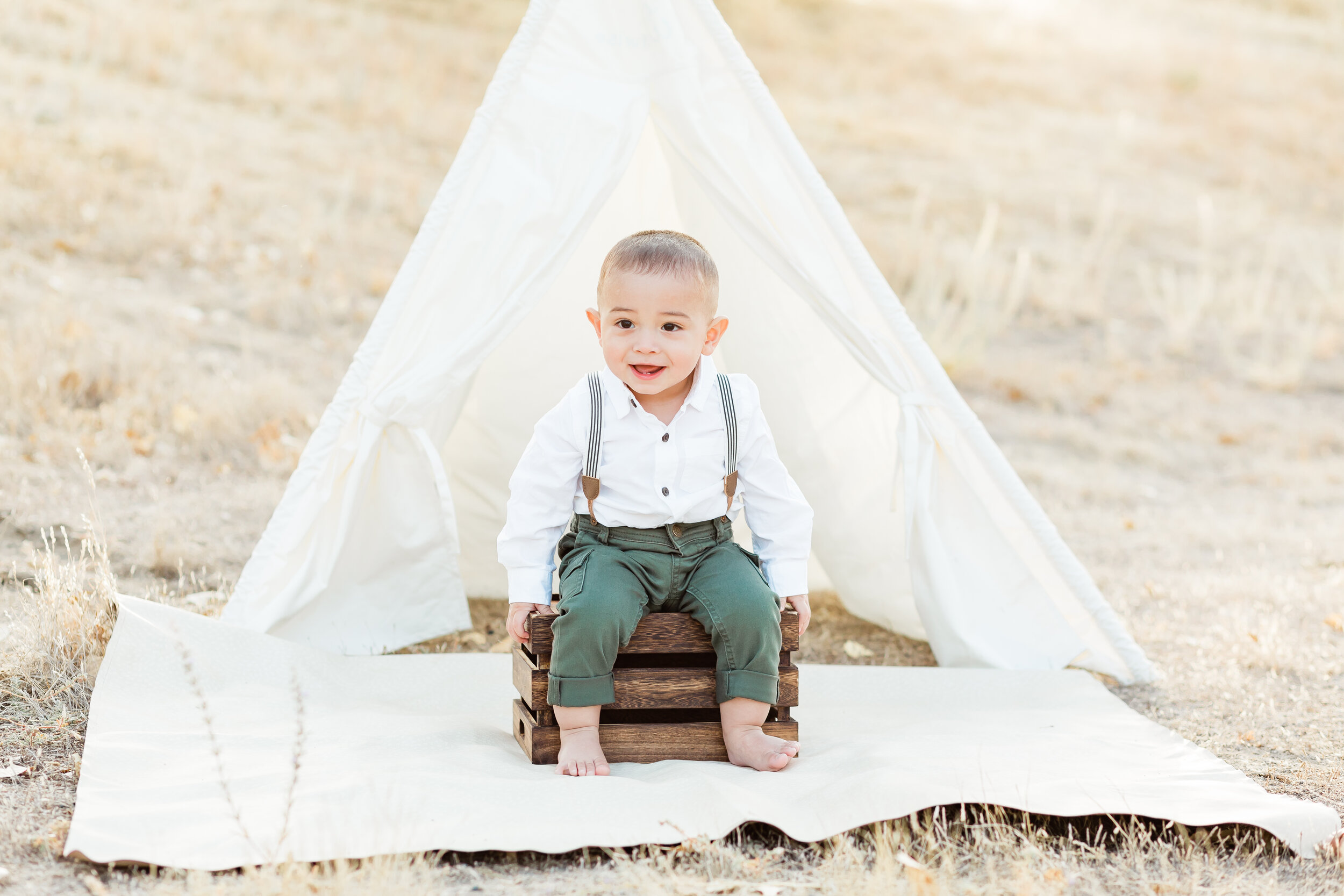 Cake Smash photo session, one year old boy wearing green pants and suspenders poses in front of a tee pee tent.