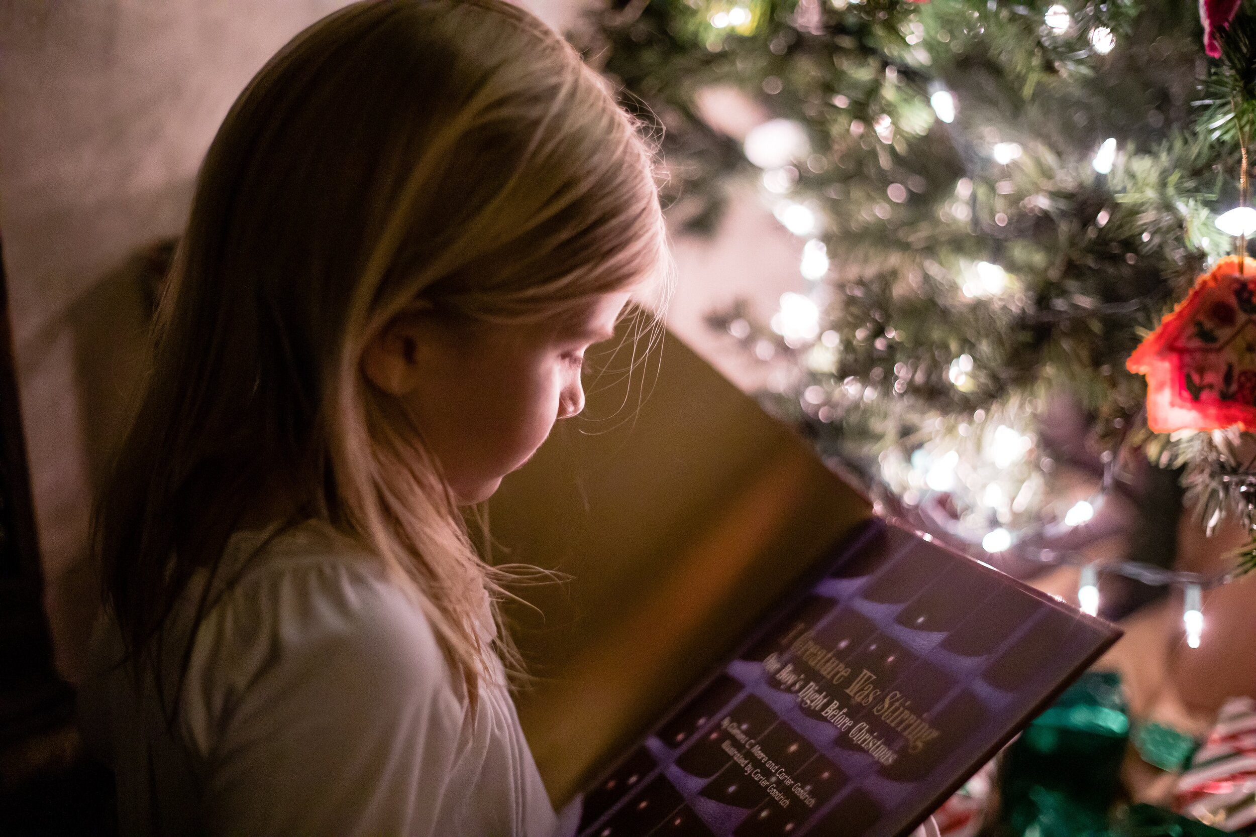 Little girl reading a book by the Christmas tree with lights on