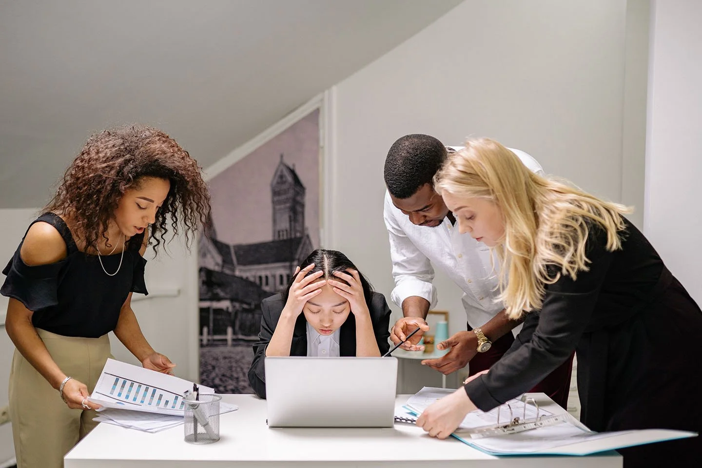 A group of four office workers, three women and one man, are gathered around a desk looking stressed while working on a laptop and reviewing documents.