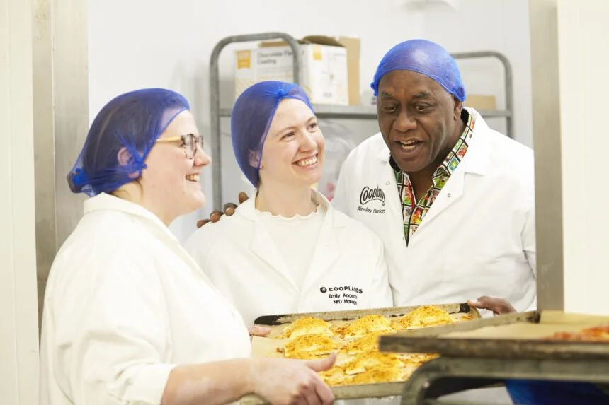 Three people from The Resilience Coach client Cooplands Bakery in white uniforms and blue hair nets smiling and looking at each other while holding a tray of freshly baked food in a commercial kitchen.