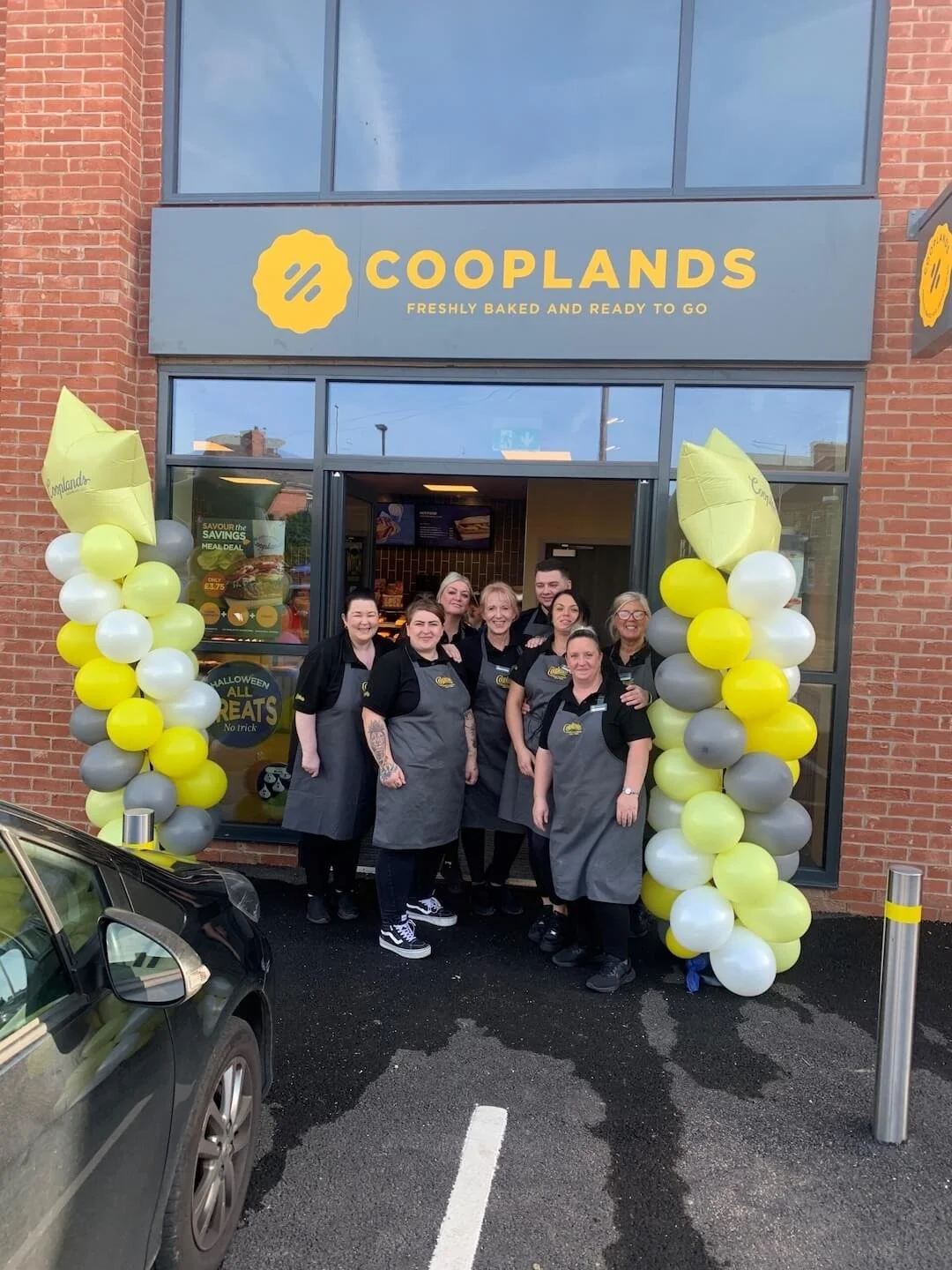Group of employees standing outside a Cooplands bakery, decorated with yellow, white, and gray balloons for a celebration or grand opening