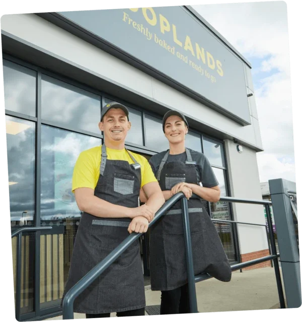 Two people, a man and a woman, stand outside in front of a Cooplands bakery store, wearing black aprons and smiling.