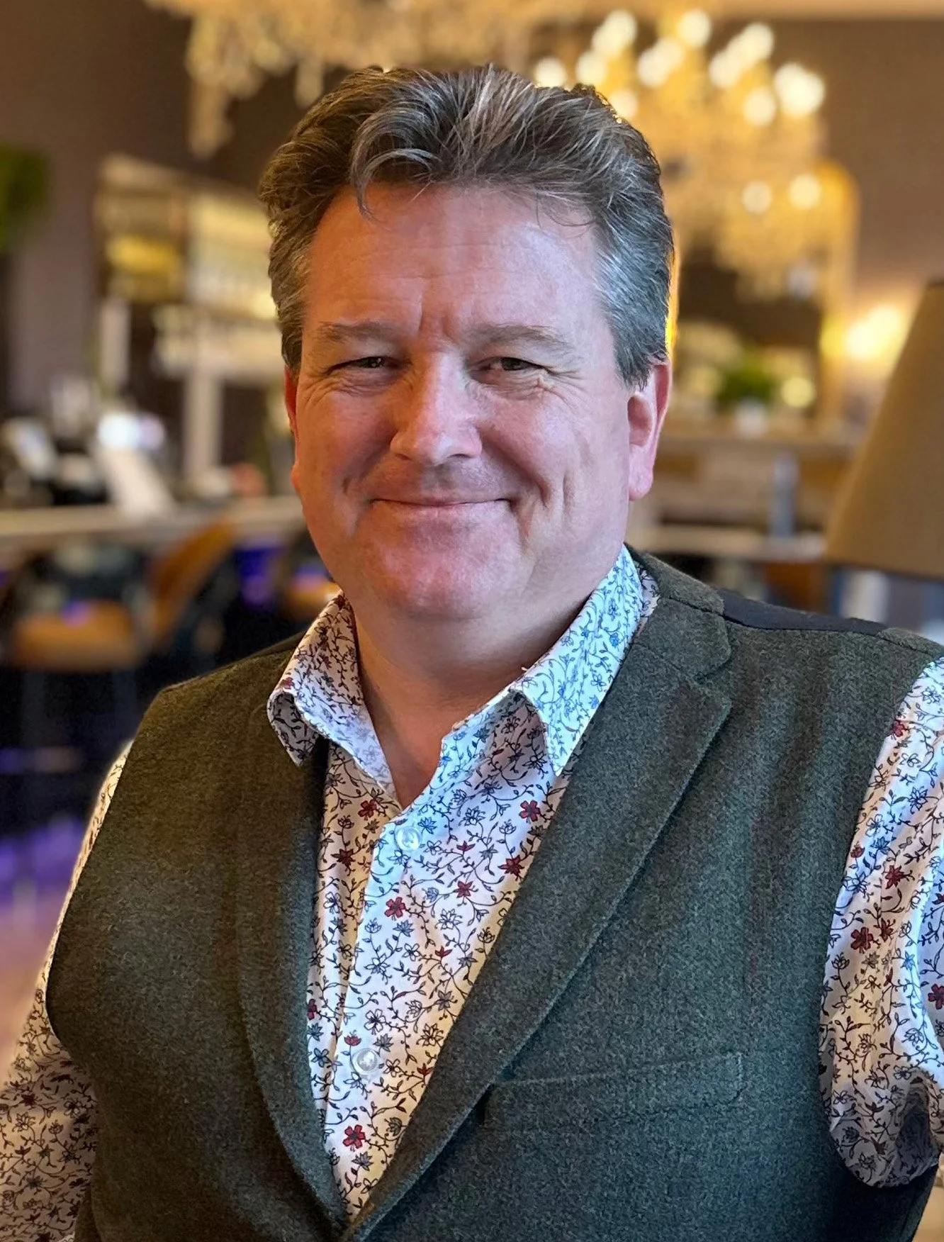 Russell Harvey with dark hair smiling, wearing a floral shirt and a dark waistcoat, sitting in a warmly lit restaurant or lounge with chandeliers and blurred background.