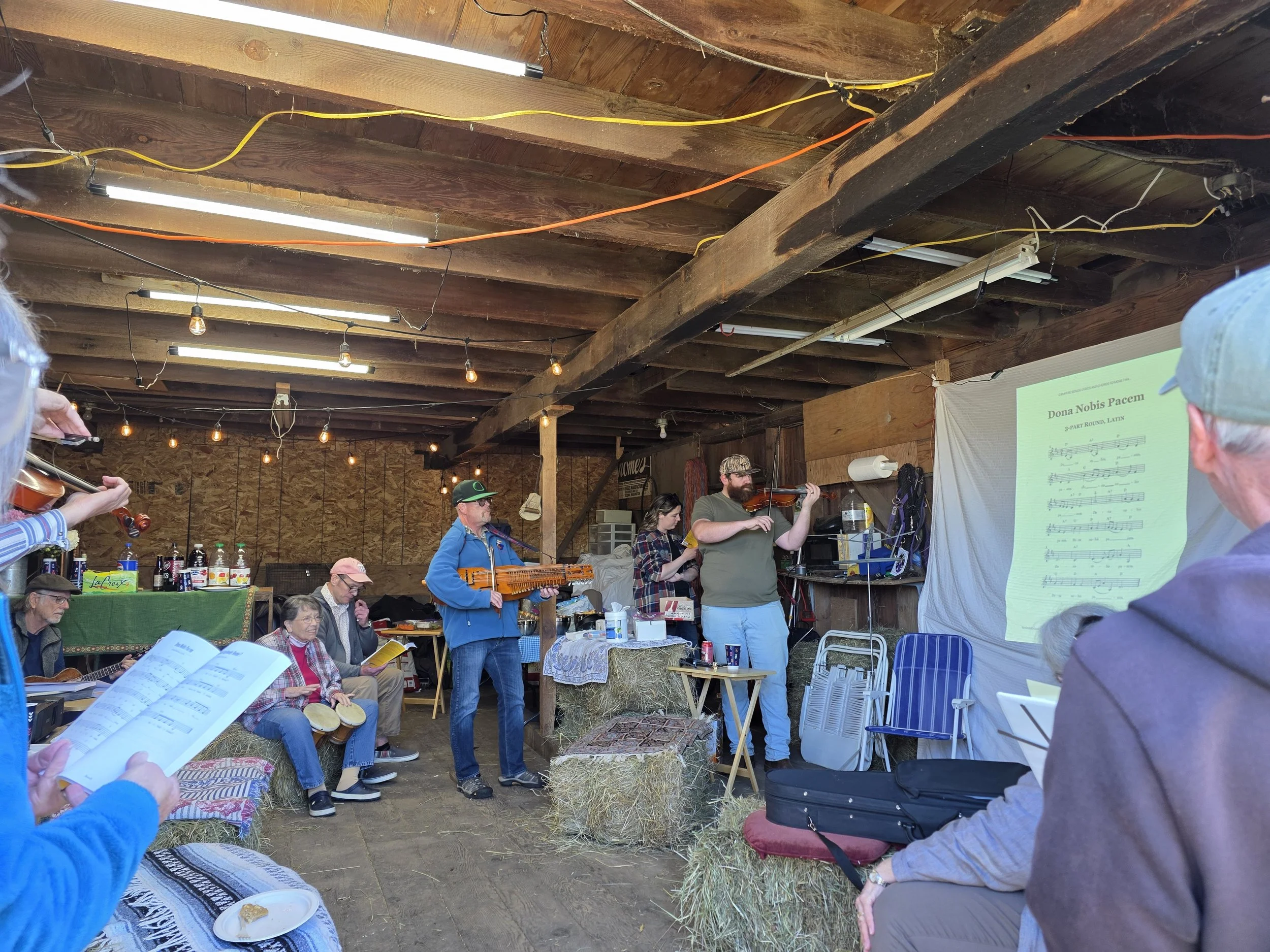 A group of people gathered inside a rustic wooden barn for a musical performance. Several musicians are playing instruments, including a violin and a hammered dulcimer, while others are reading sheet music. String lights are hanging from the ceiling, and a projector screen displaying sheet music is on the right side. Some people are seated on hay bales, and a table with snacks and drinks is in the background.