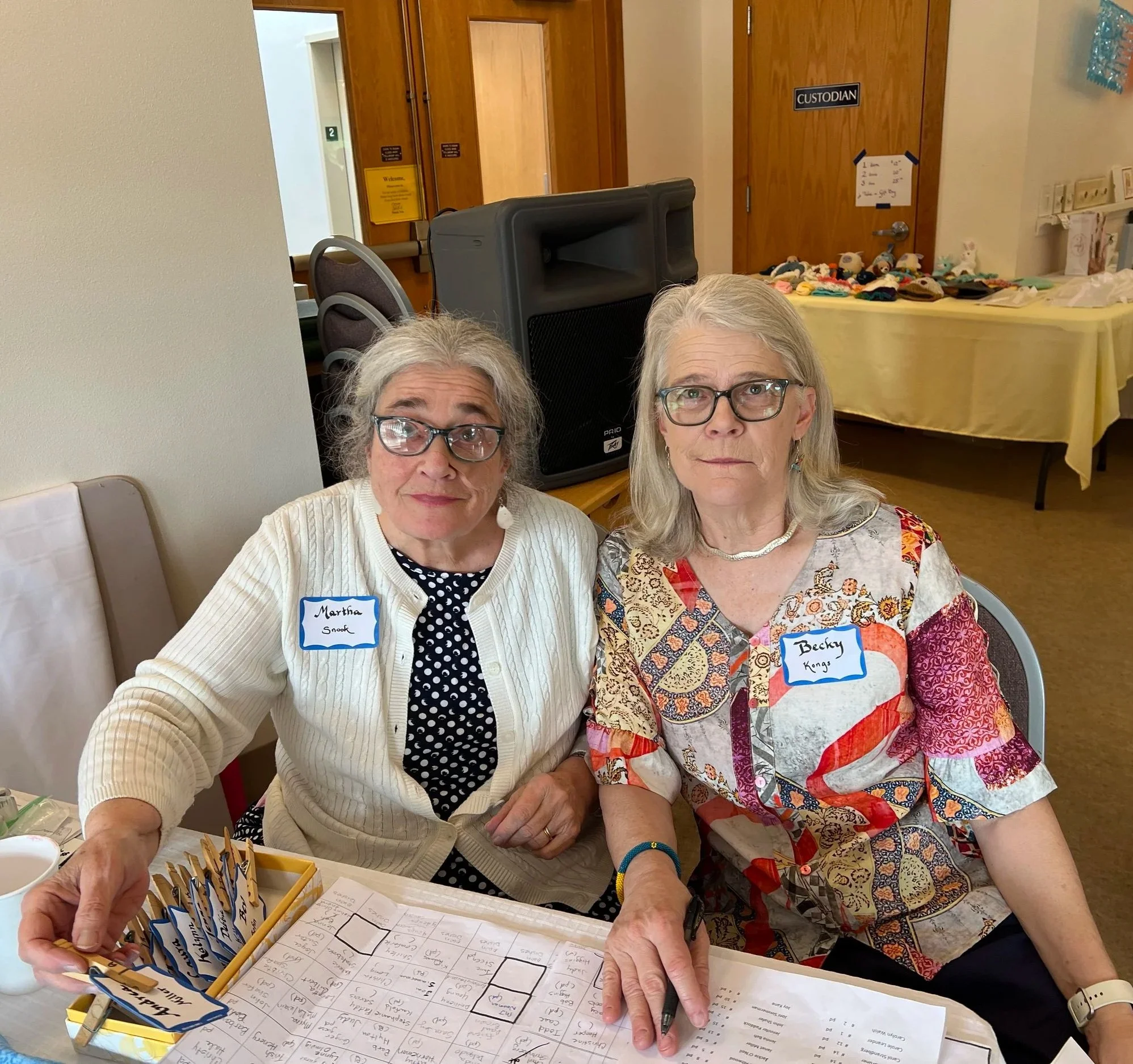 Two elderly women with glasses sitting at a table during a gathering or event. One woman is wearing a white cardigan and has a name tag that reads Martha Snook, while the other woman is wearing a colorful patterned blouse and has a name tag that reads Becky. They are looking towards the camera. On the table, there are name tags, a sheet of paper, and pens. In the background, there is a door labeled 'CUSTODIAN' and a table with colorful items and signs.
