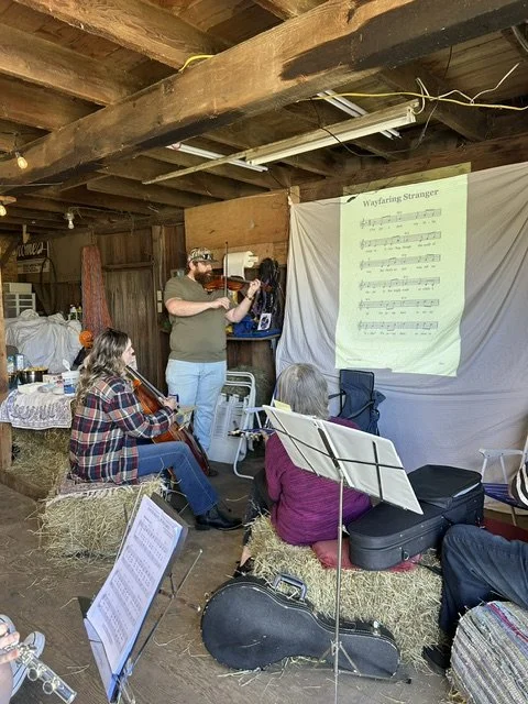 A small group of people gathered in a rustic barn, with some sitting on hay bales, watching a person play the violin, while a music sheet titled "Wayfaring Stranger" is projected on a white sheet on the wall.