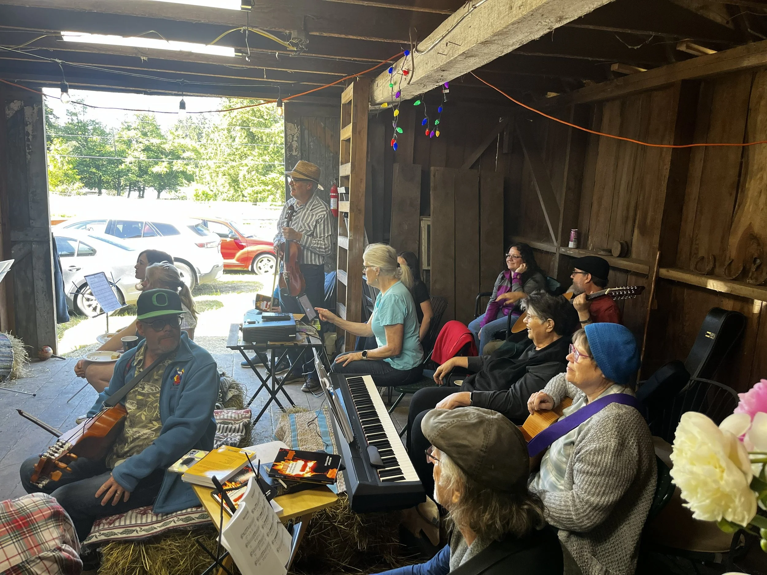 A group of people gathered in a rustic barn with wooden walls and hanging string lights, performing or rehearsing music with guitars, a keyboard, and sheet music, during the daytime.