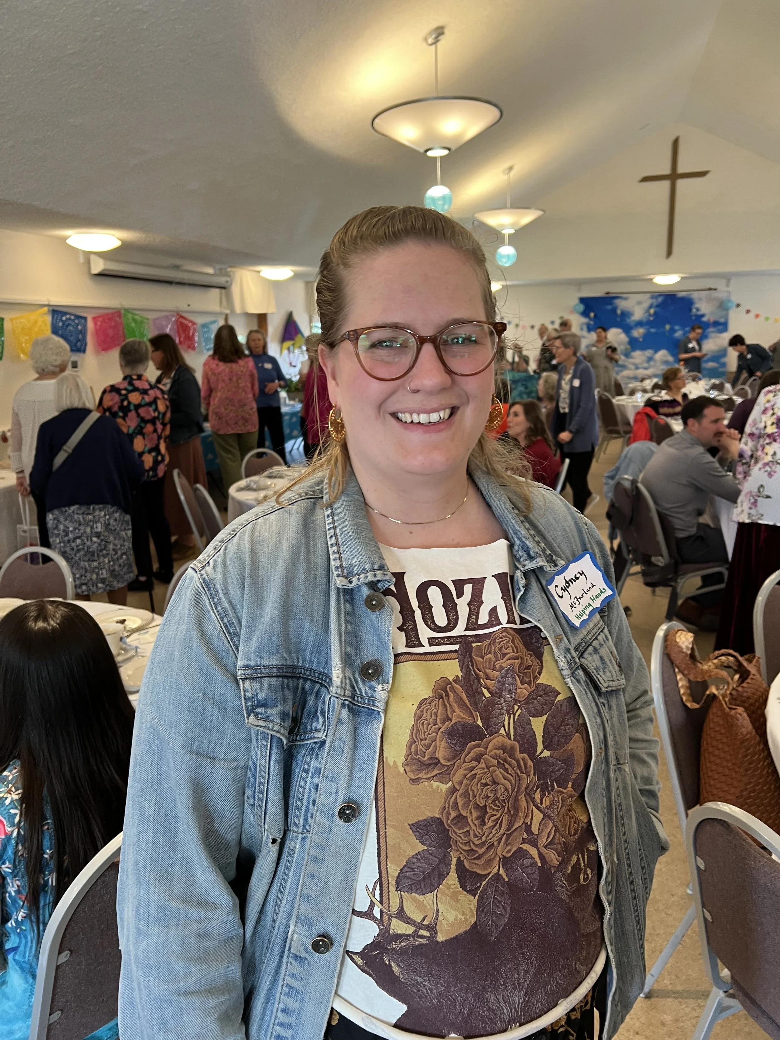 A woman wearing glasses, a denim jacket, and a floral shirt standing in a decorated hall with many people, tables, and festive decorations.