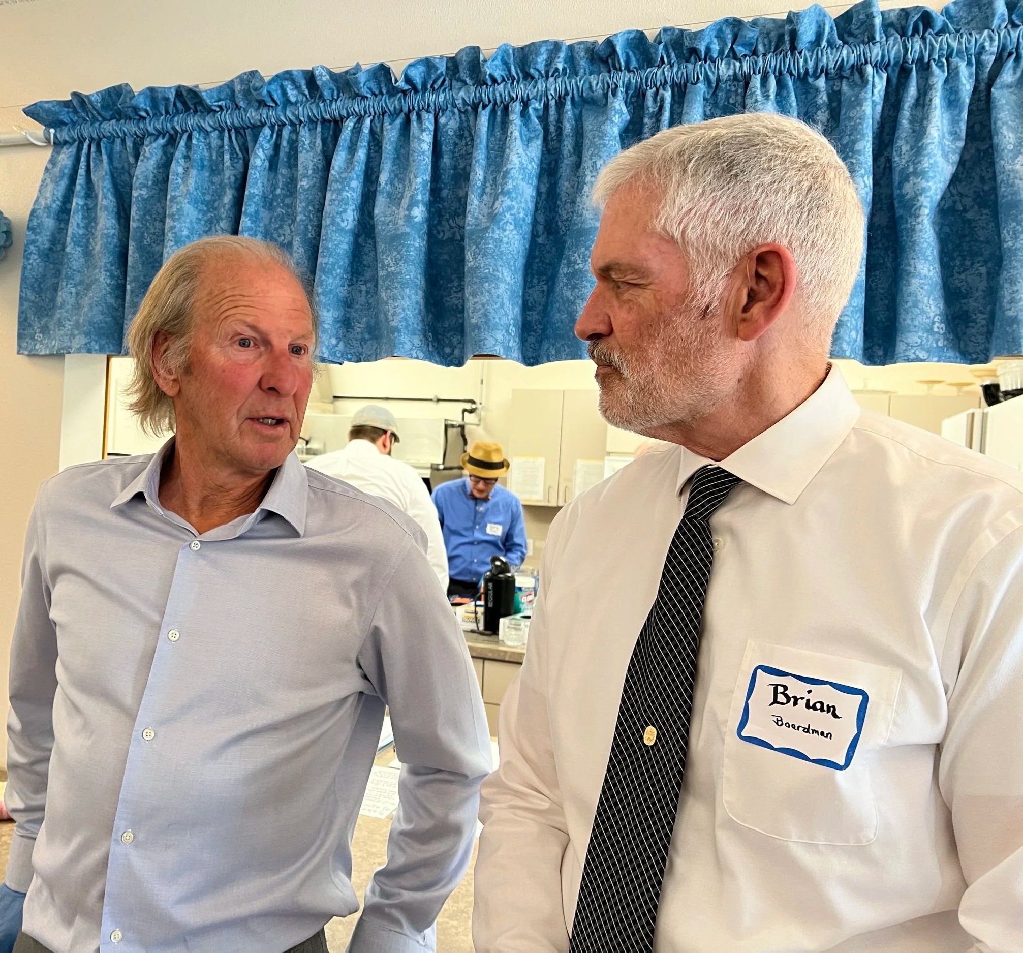 Two men engaging in conversation in an indoor setting, with a woman wearing a hat in the background and a blue curtain overhead.