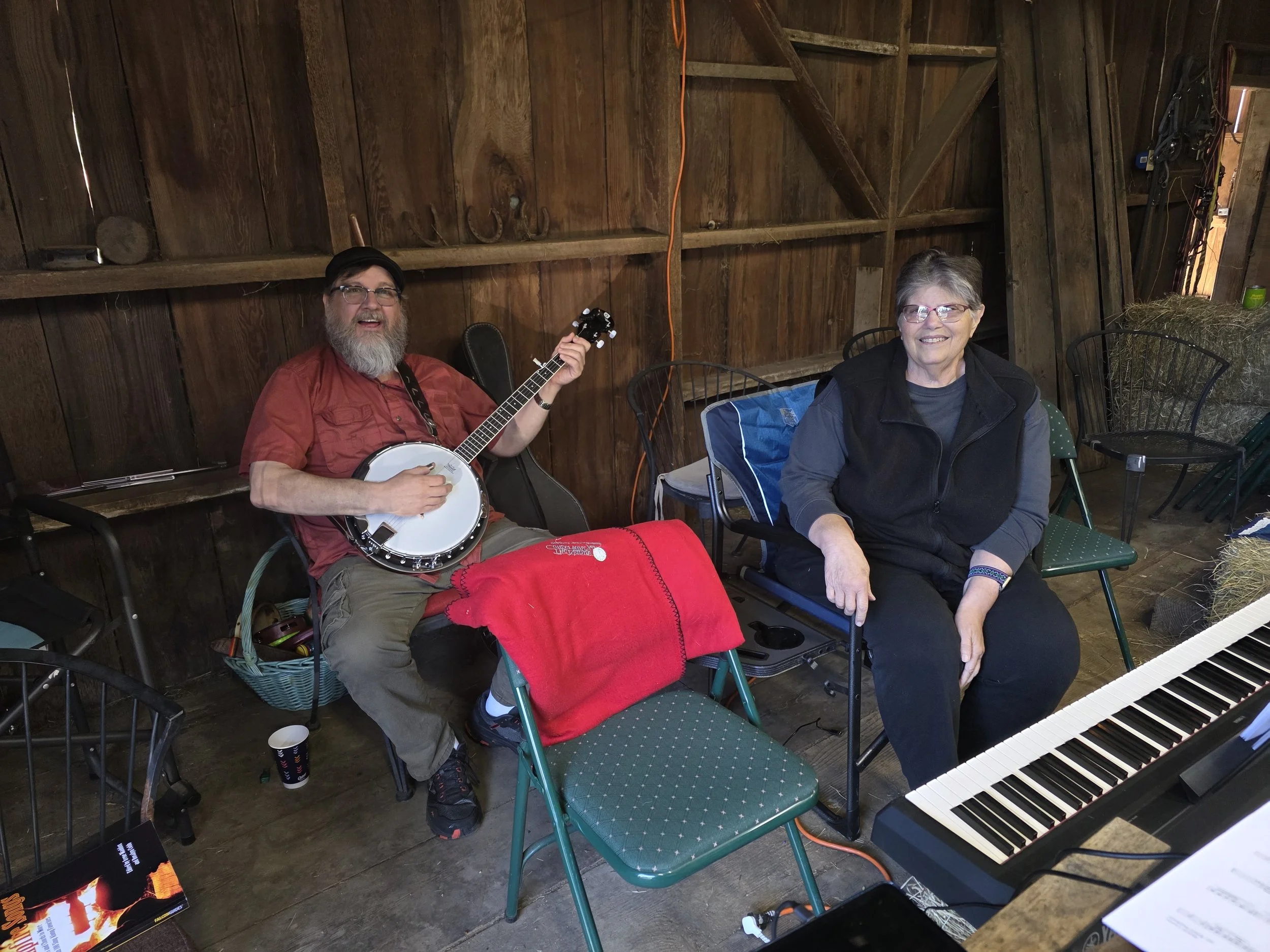 Two people are seated in a wooden room. The man on the left has a beard, glasses, and is playing a banjo. The woman on the right has short gray hair, glasses, and is smiling. There is a keyboard in front of them and a book on the floor nearby.