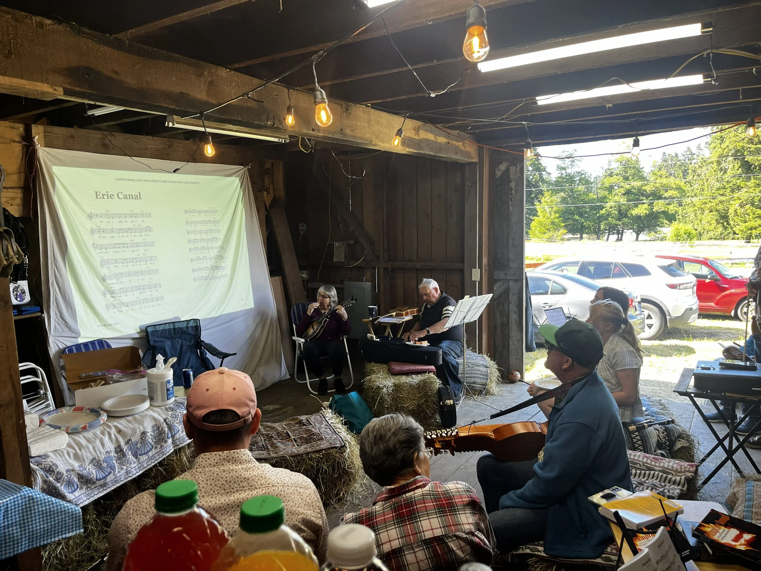 Indoor gathering of people seated on hay bales and chairs, playing guitars and music, with a large sheet displaying sheet music on a wall, in a rustic barn with string lights and large open door showing parked cars and trees outside.
