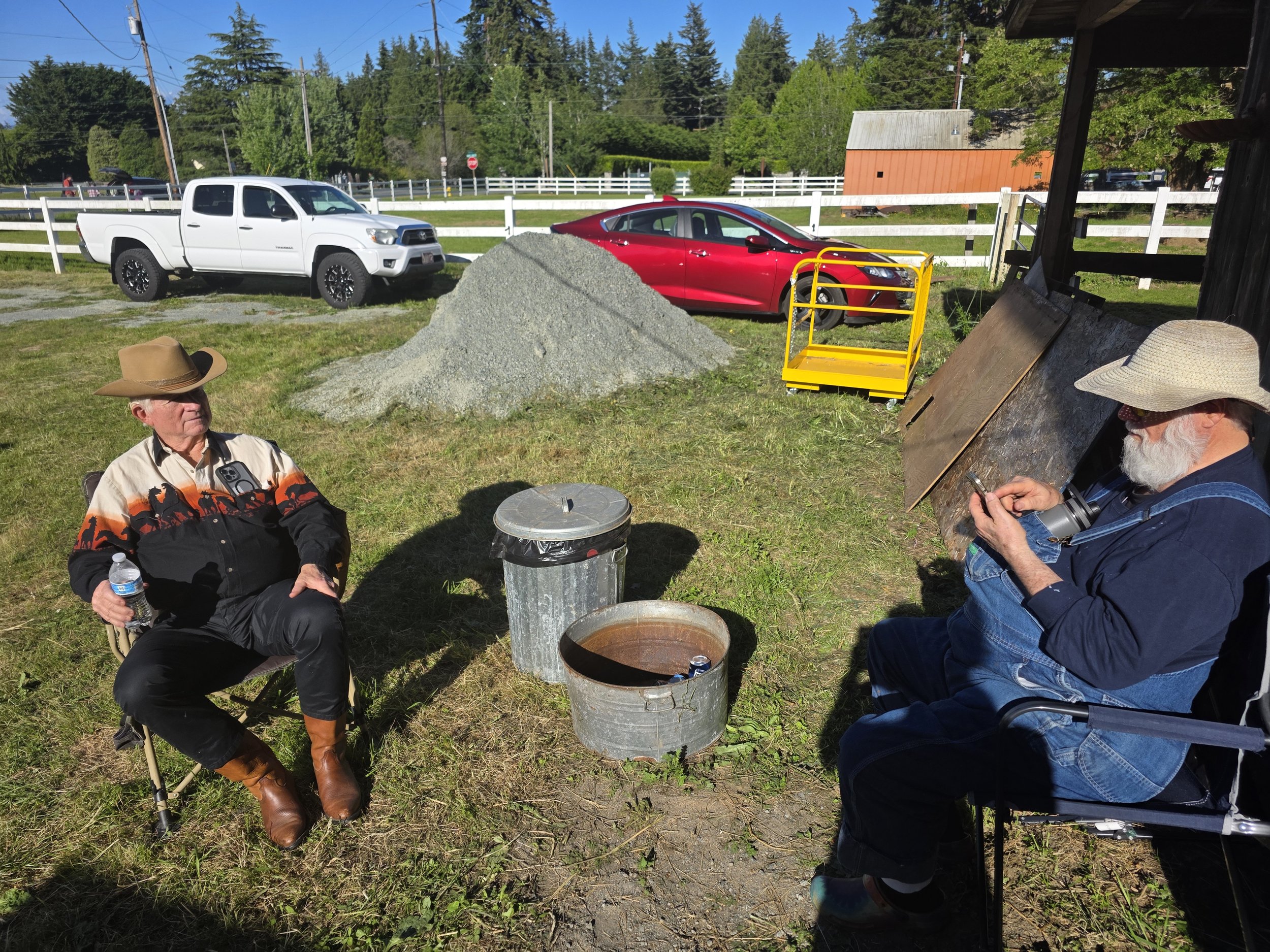 People sitting outdoors in a sunny area with a white truck and red car behind them. There is a pile of gravel, a yellow cart, and a large trash can nearby. The background has trees, a white fence, and a small orange building.