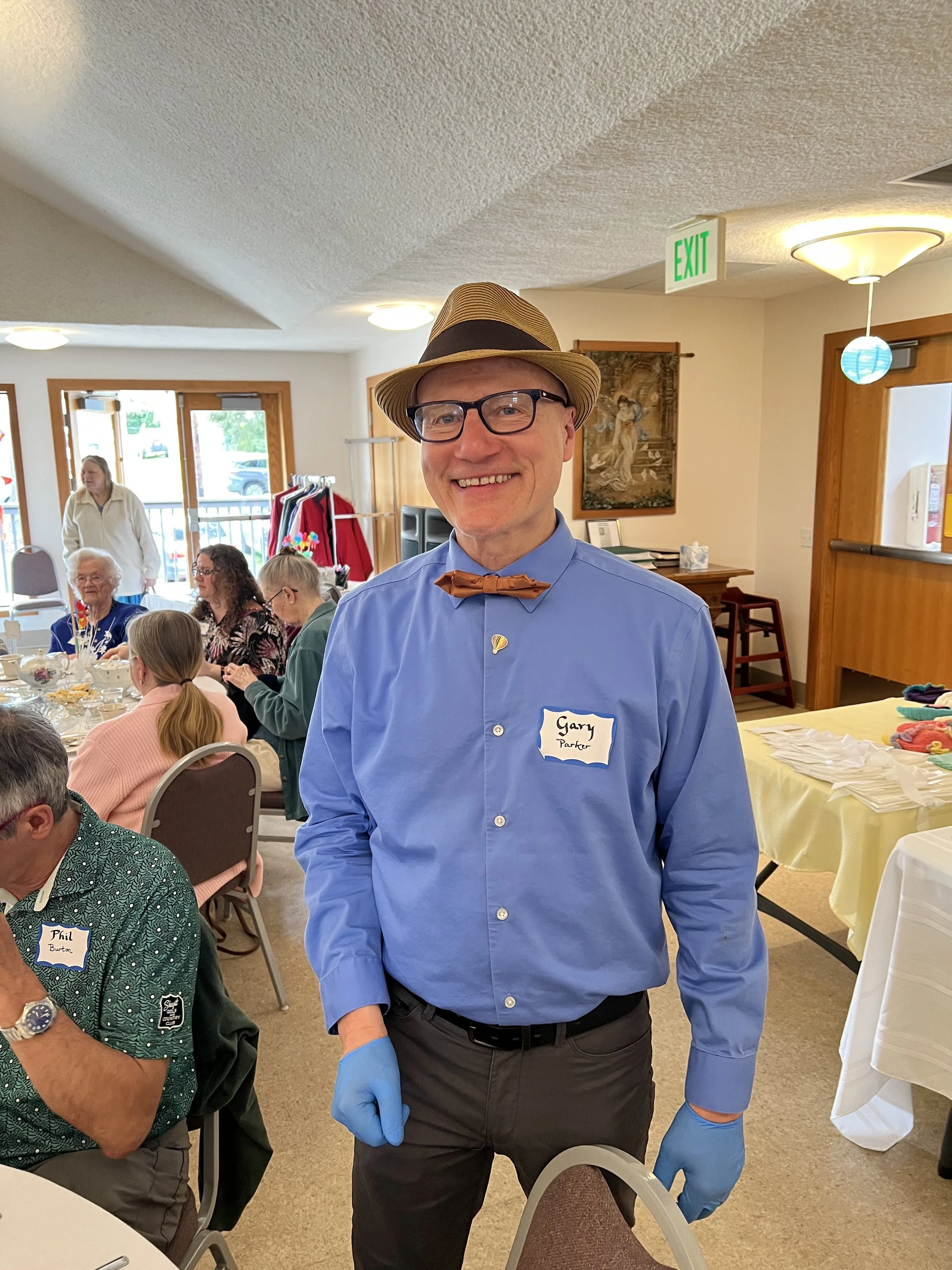 A man wearing a light blue dress shirt, black pants, a brown bow tie, a beige fedora with a black band, black glasses, and blue gloves, smiling at camera at a social gathering or party indoors with other guests seated at tables, some wearing name tags.