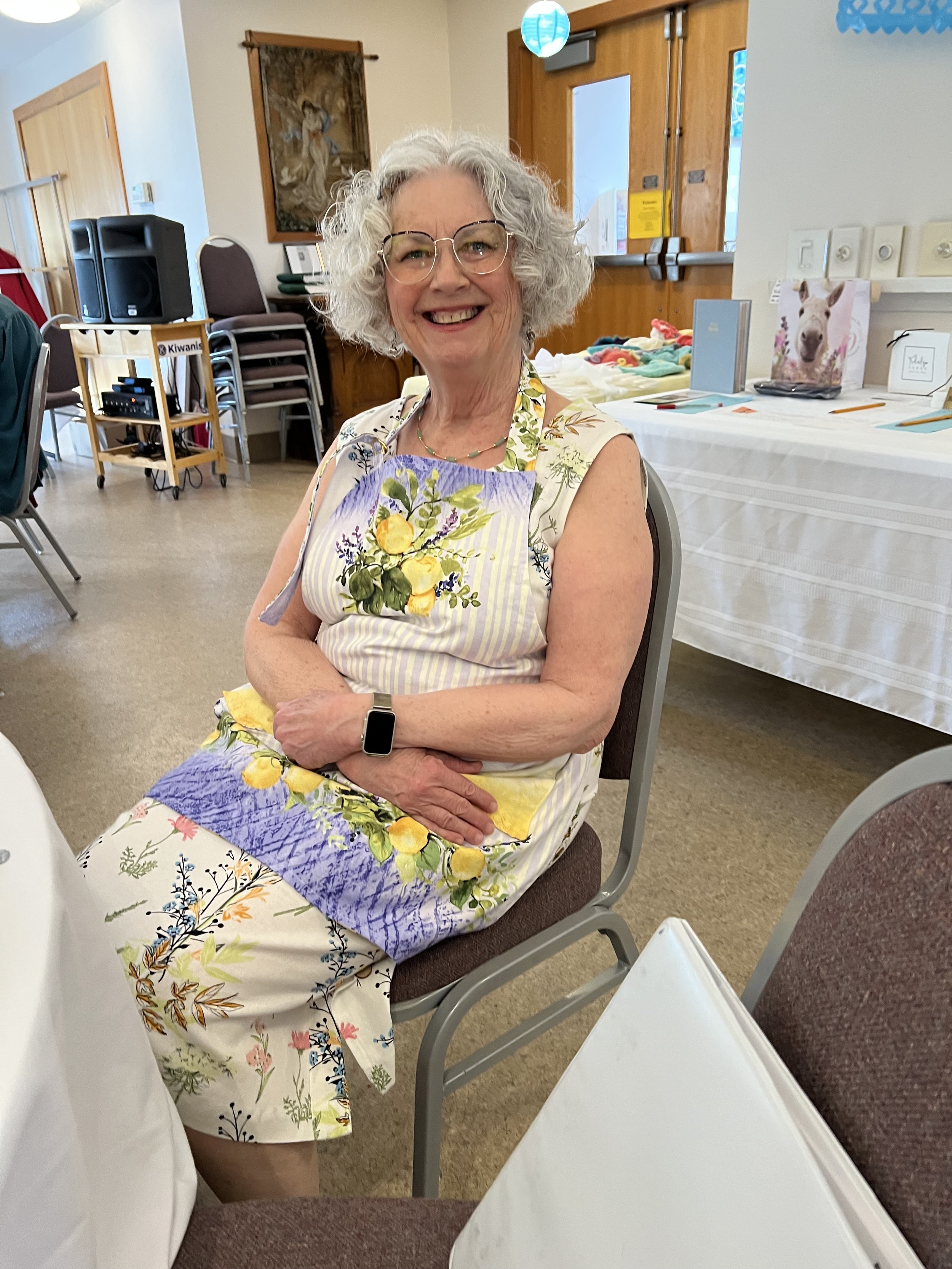 A smiling older woman with curly gray hair and glasses sitting at a table in a decorated room. She is wearing a floral apron with lemons and other plants on it, and a sleeveless top. Behind her are chairs, a cart, and a table with colorful items, possibly at a gathering or event.