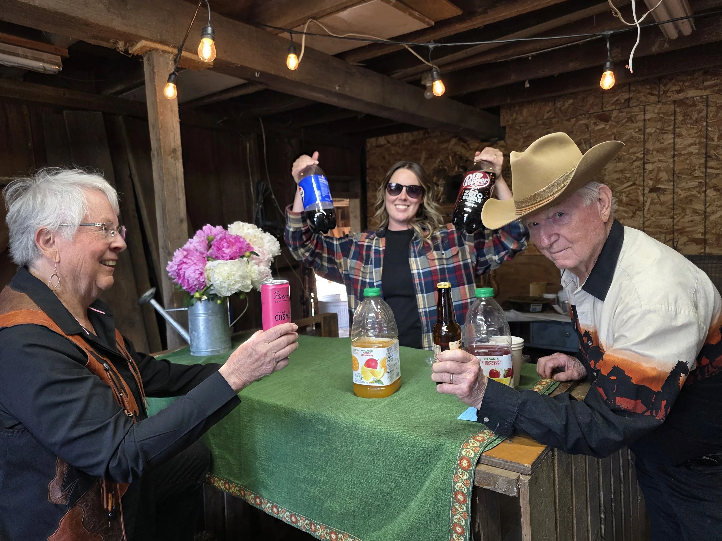 Three women and one man are sitting at a table in a rustic setting, with one woman in the middle holding two bottles of soda and smiling. The table has a green tablecloth, a bouquet of flowers, and various drink bottles. The surroundings include wooden walls and exposed beams, with hanging light bulbs overhead.