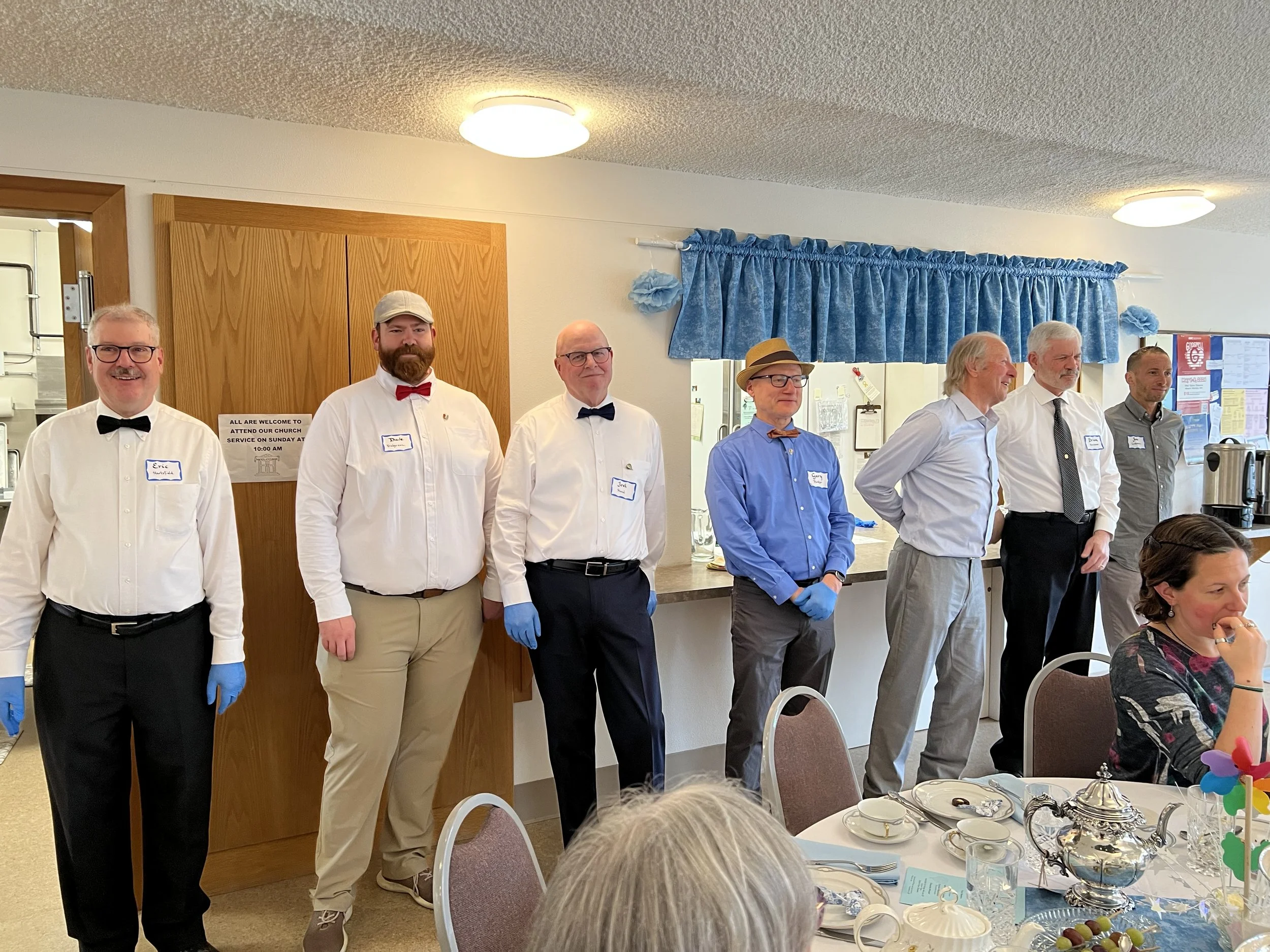 Group of men standing in a line at a banquet table, dressed in semi-formal attire with bow ties, some wearing gloves. A woman sitting at the table is visible in the foreground.