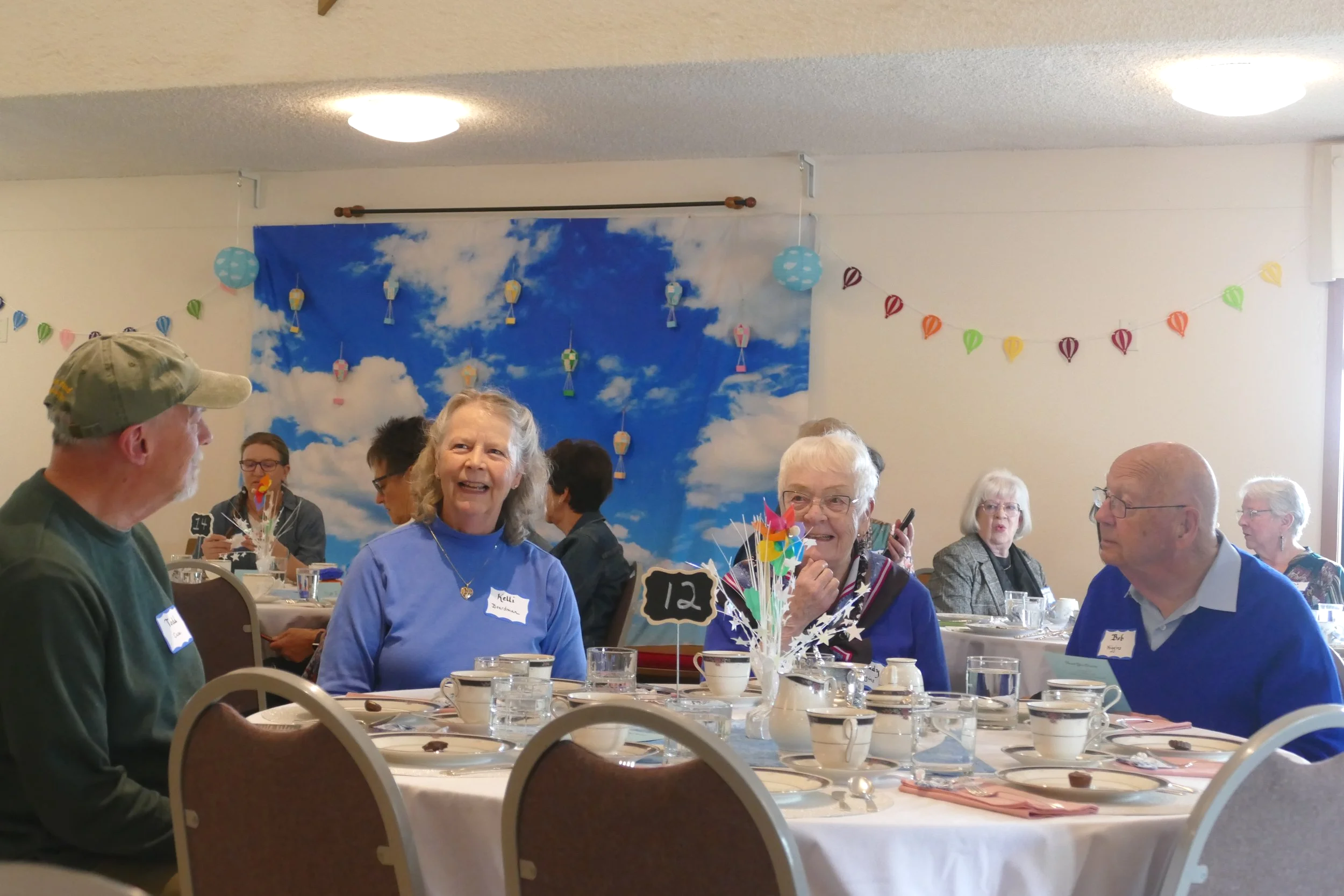 A group of elderly people sitting at a long banquet table during a celebration or gathering, decorated with colorful lanterns and a blue sky-themed backdrop with clouds.