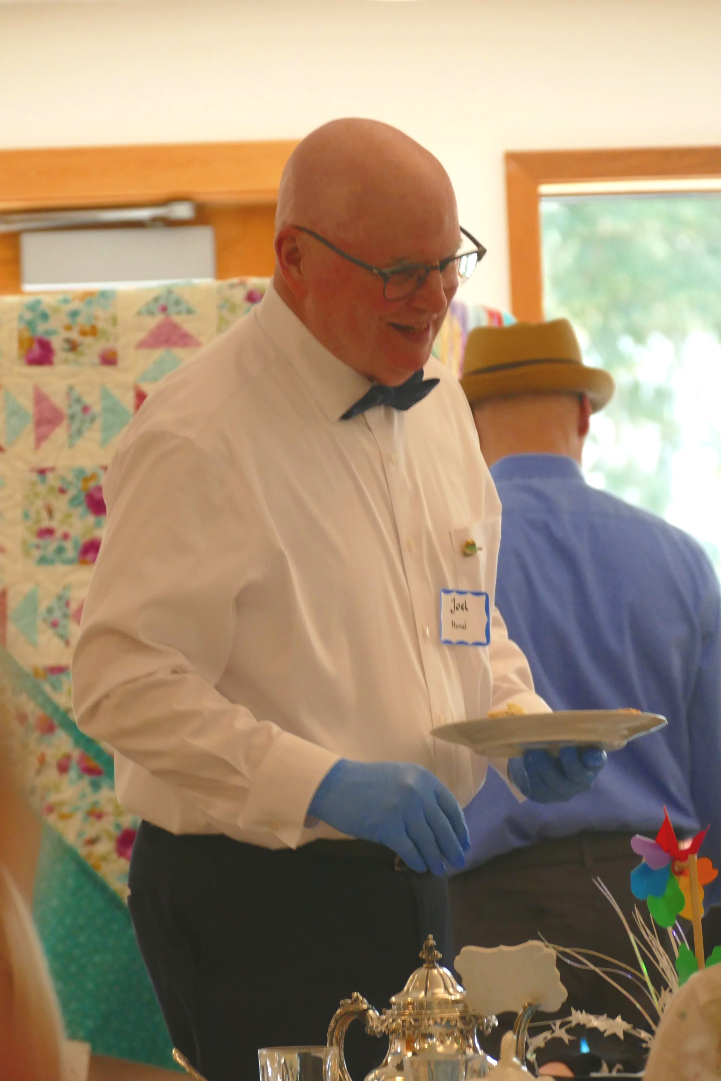 A bald man wearing glasses, a white dress shirt, a black bow tie, and blue gloves holding a silver tray, smiling at an indoor event. In the background, there is a quilted fabric with colorful triangles, a person in a straw hat, and a window with green trees outside.
