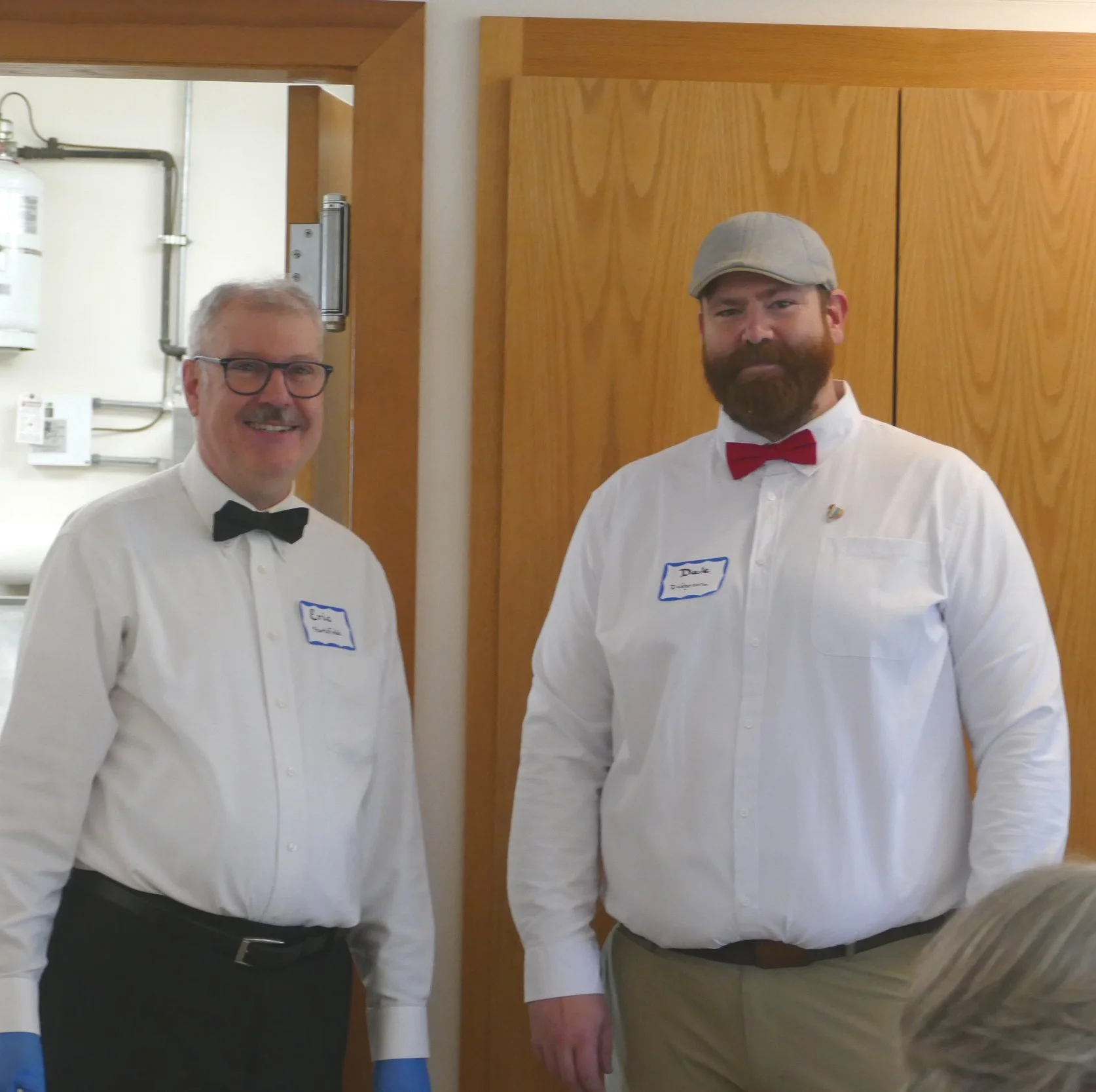 Two men dressed in white shirts, one with glasses and a black bow tie, the other with a gray cap and red bow tie, standing together indoors against a wooden wall.