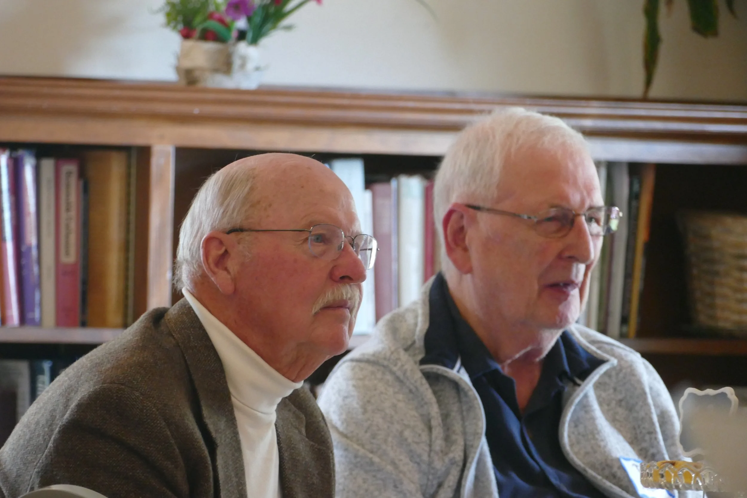 Two elderly men with glasses and light-colored hair sitting side by side indoors with bookshelves and a potted plant in the background.