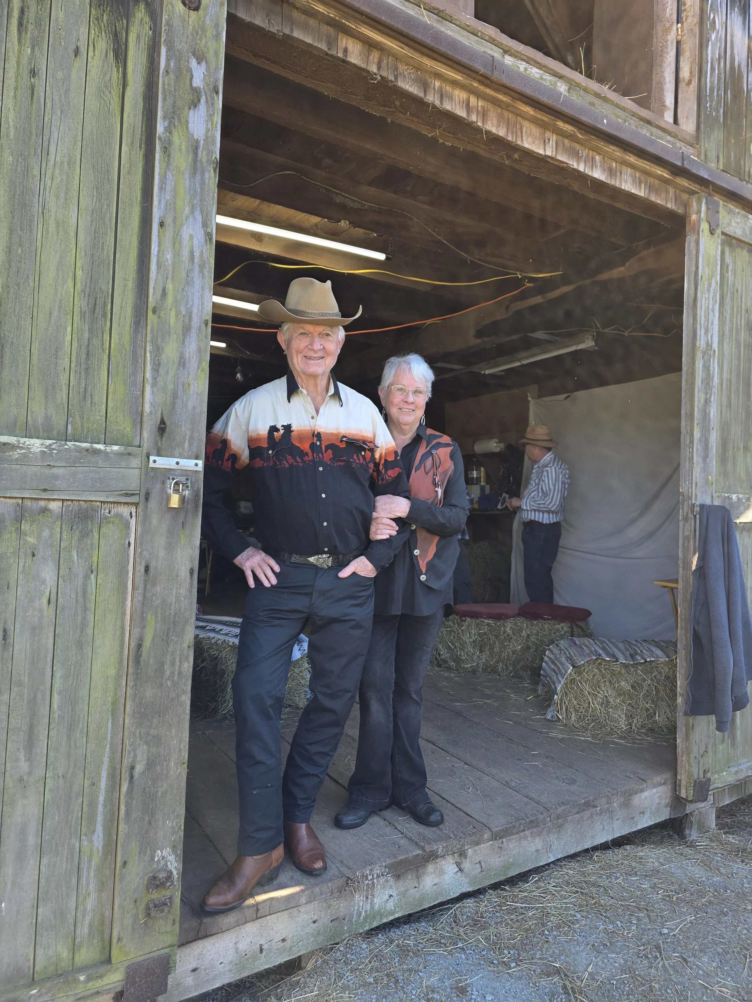 An older man and woman standing inside a rustic barn with hay bales, looking at the camera. The man wears a cowboy hat and western shirt, and the woman is in a southwestern style jacket with earrings. In the background, a man in a hat is working near a counter.