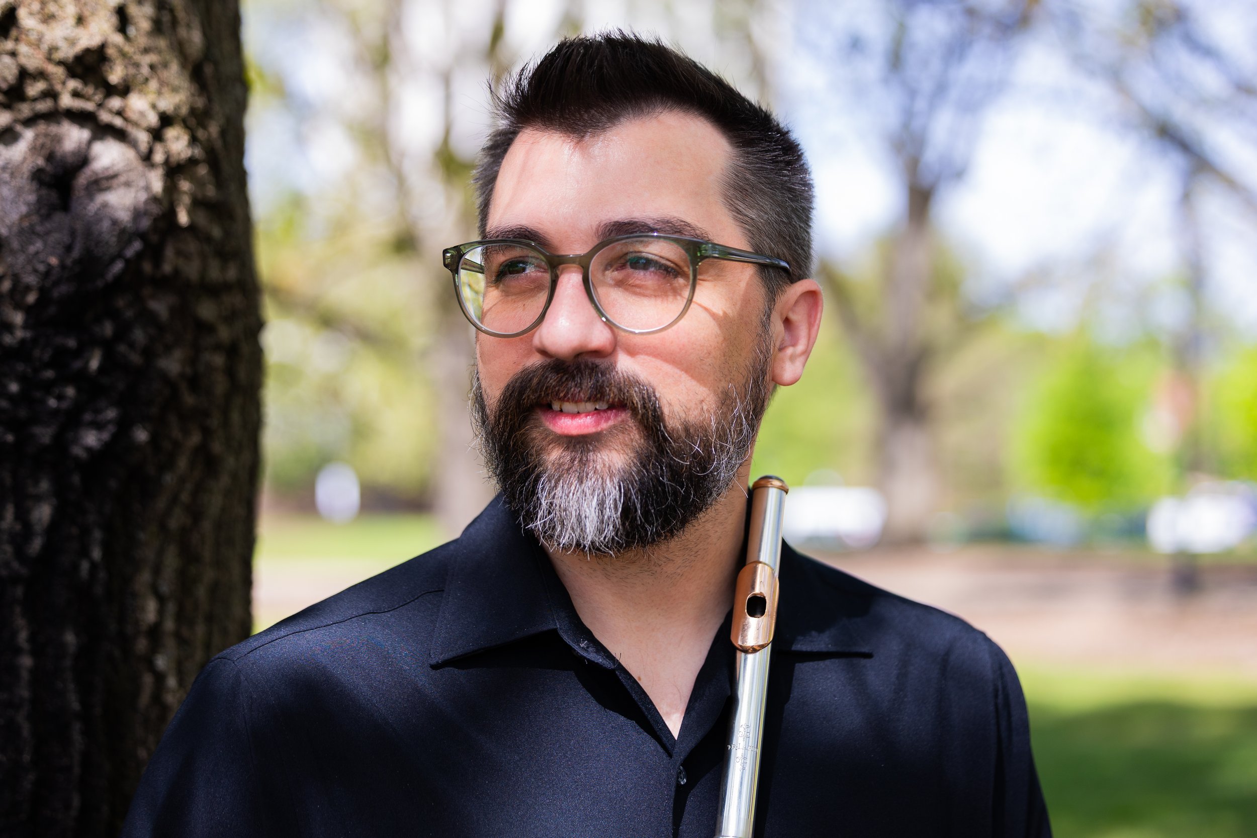 Man with eye glasses a black shirt stands in front of a tree while holding a flute.