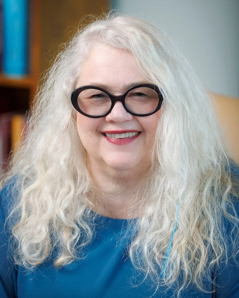 A woman with long blonde hair wearing round black glasses and black lipstick, smiling.