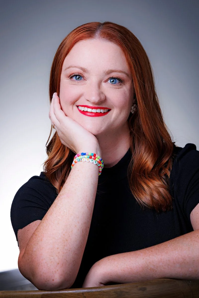 A smiling woman with red hair and blue eyes, wearing a black shirt, resting her chin on her hand, with colorful bead bracelets on her wrist.
