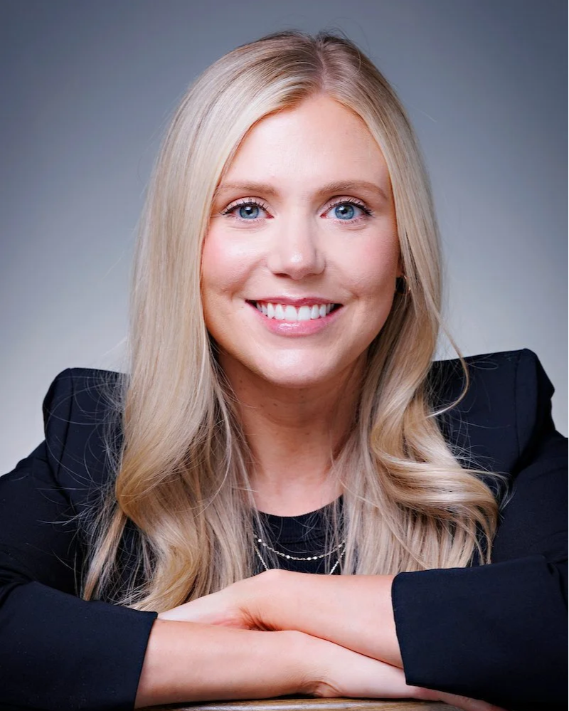 A young blonde woman with bright blue eyes smiling at the camera, touching her face with her left hand, wearing a coral-colored top, standing in front of a plain wall with a wooden object in the background.
