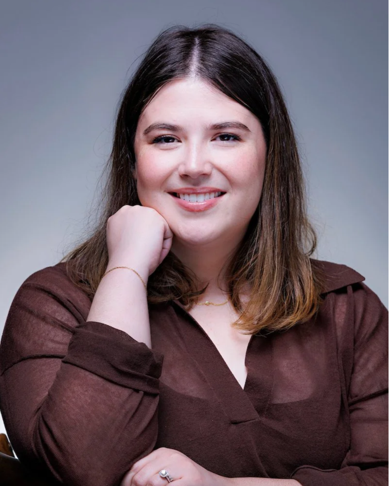 A woman with long brown hair, smiling and resting her chin on her hand, sitting at a table against a light-colored background.
