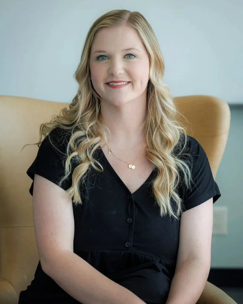 Close-up portrait of a woman with long wavy blonde hair, blue eyes, pink lipstick, wearing a gray ribbed sweater, gold jewelry, standing in front of a bookshelf with colorful books.