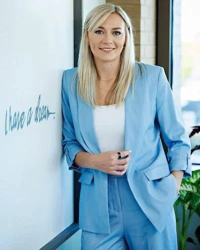 A woman with long blonde hair wearing a light blue suit and white shirt, standing next to a whiteboard with partially visible text, in a modern office setting.