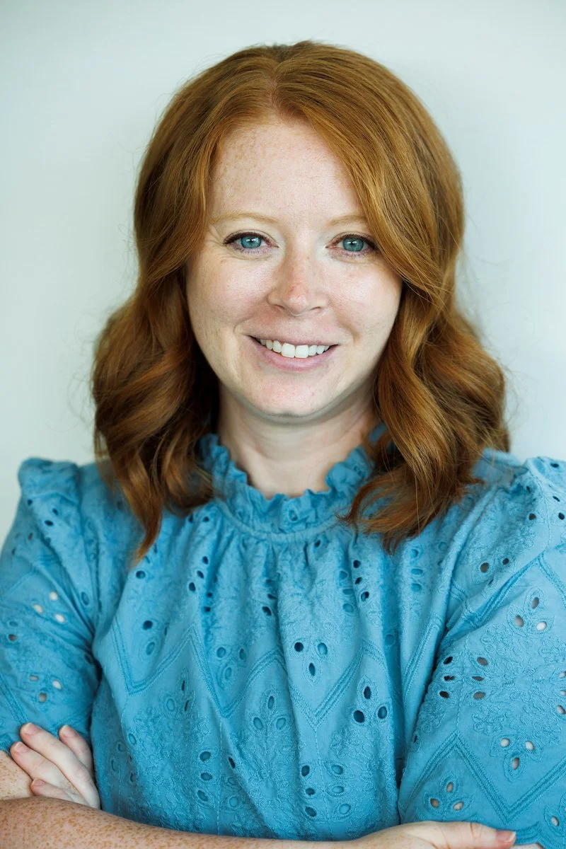 A woman with red hair and blue eyes smiling, wearing a blue eyelet blouse, standing against a light background.