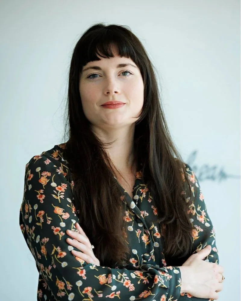 A woman with long dark hair, wearing a black floral blouse, standing with arms crossed against a neutral background.