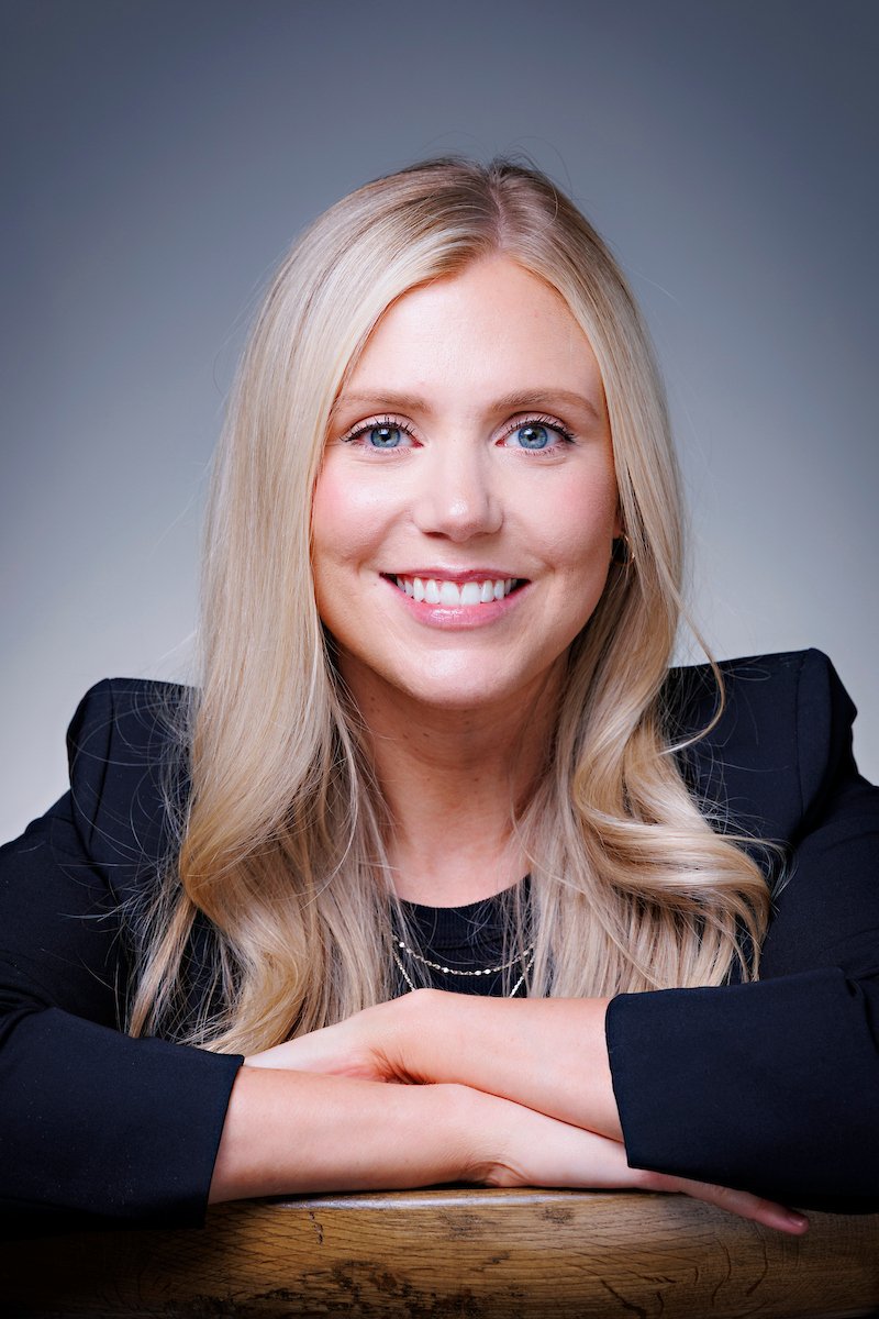 Close-up portrait of a smiling blonde woman with blue eyes, wearing a black blazer, resting her arms on a wooden surface against a blurred gray background.