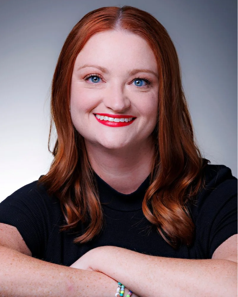 Close-up portrait of a smiling woman with red hair, blue eyes, and wearing a blue scarf and top, against a white background.