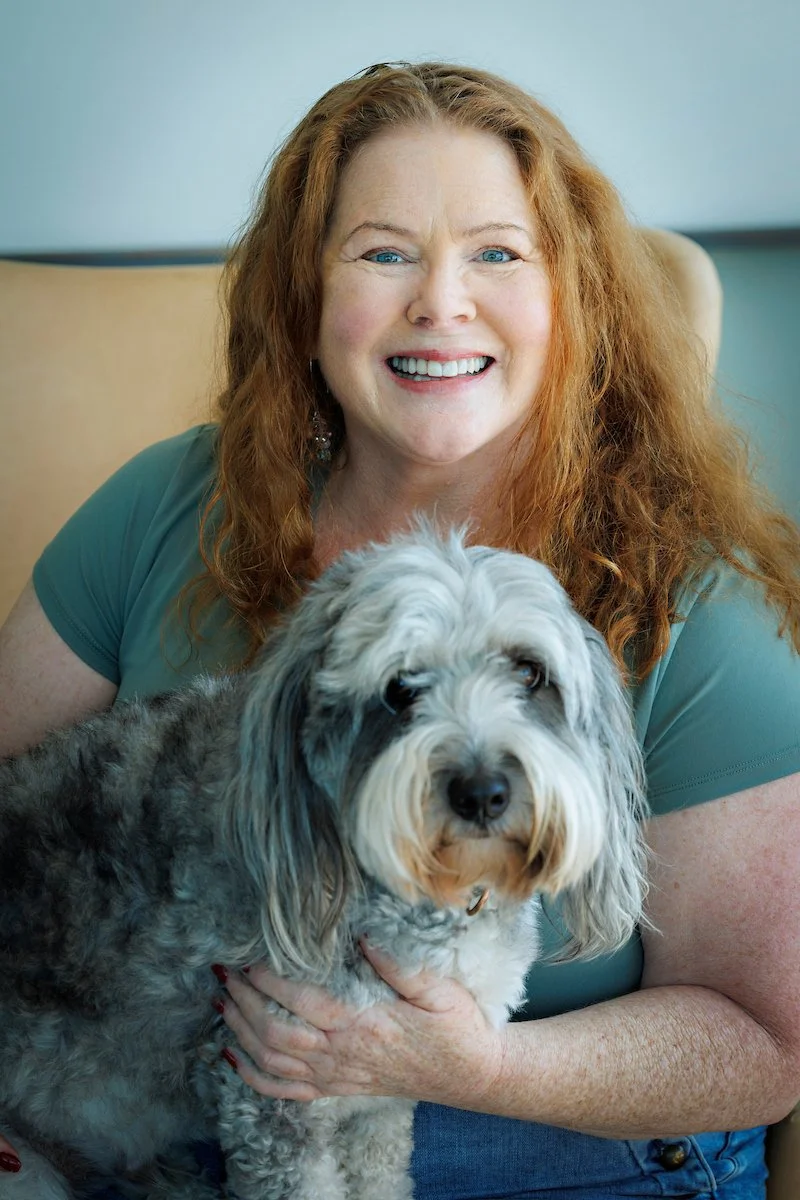 A woman with curly red hair smiling and holding a black and white dog with long fur, sitting on a beige chair in a room with light blue walls.