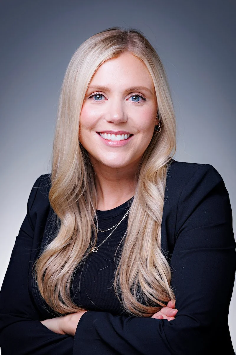 A smiling blonde woman with blue eyes and a black top, smiling with arms crossed, against a neutral gray background.