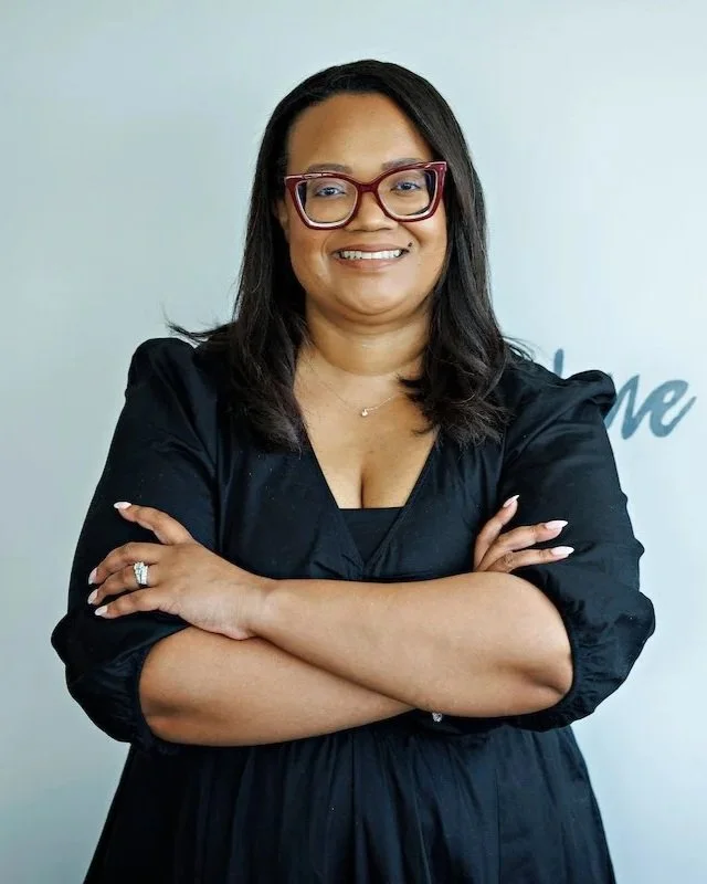 Close-up of a woman with shoulder-length black hair, wearing red glasses, a purple blouse, and jewelry, standing with her arms crossed and smiling against a blurred background.