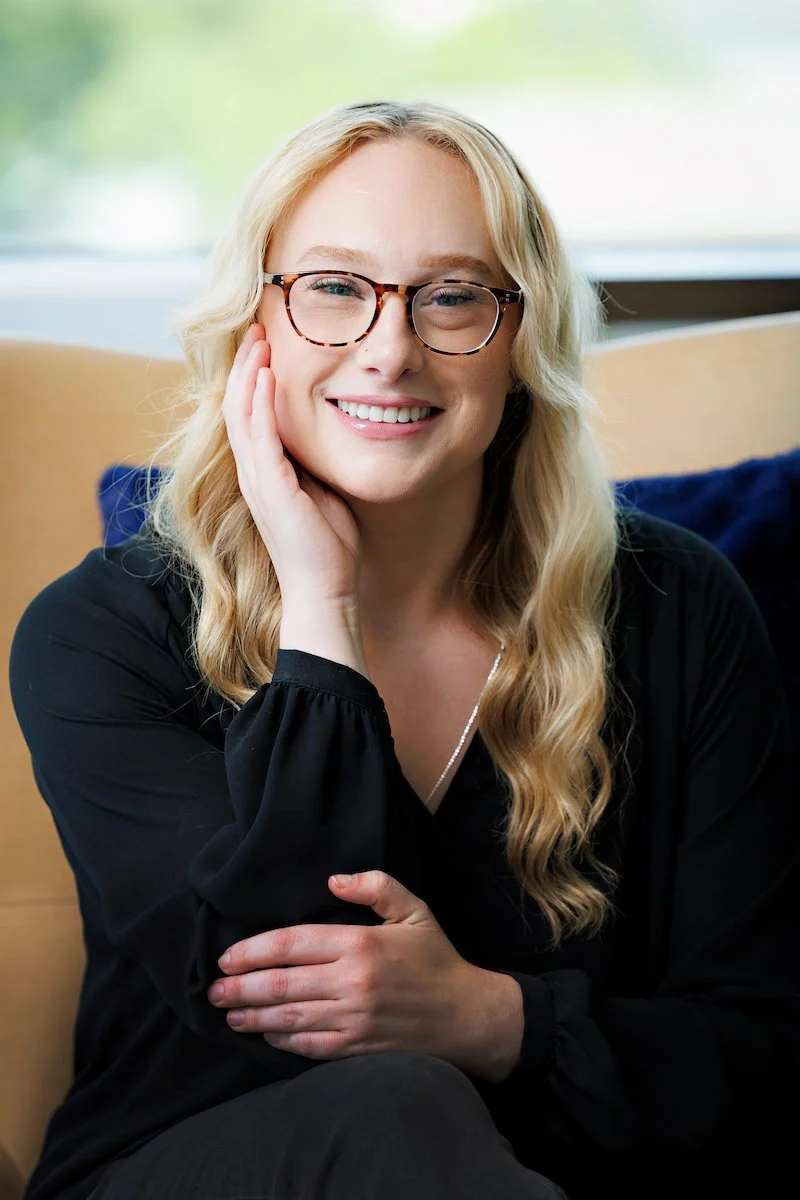A young woman with long, blonde, curly hair smiling at the camera, wearing a black top and a necklace, with a softly lit background including a lamp.