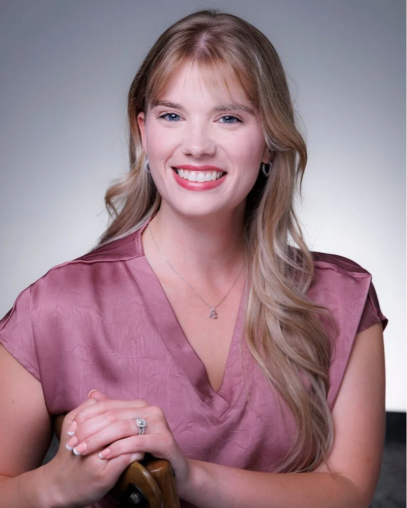 A woman with long blonde hair, blue eyes, and light skin, smiling, wearing a mauve blouse and jewelry, and holding her hands together, against a neutral background.