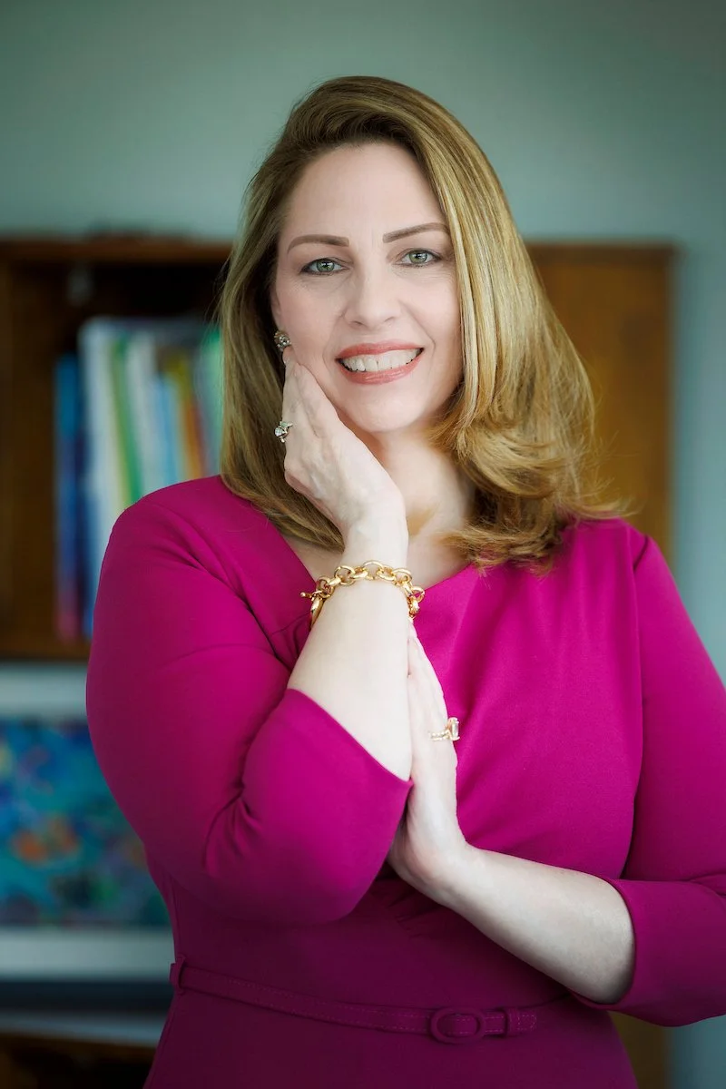A woman with shoulder-length light brown hair, wearing a fuchsia dress, smiling and gently touching her face with her right hand, in a professional indoor setting with a bookshelf in the background.