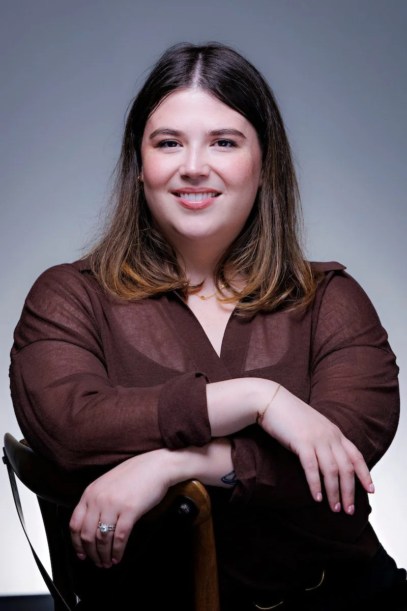 A woman with shoulder-length brown hair smiling at the camera, wearing a brown blouse with rolled-up sleeves, sitting with one arm resting on a wooden chair armrest, against a gray background.
