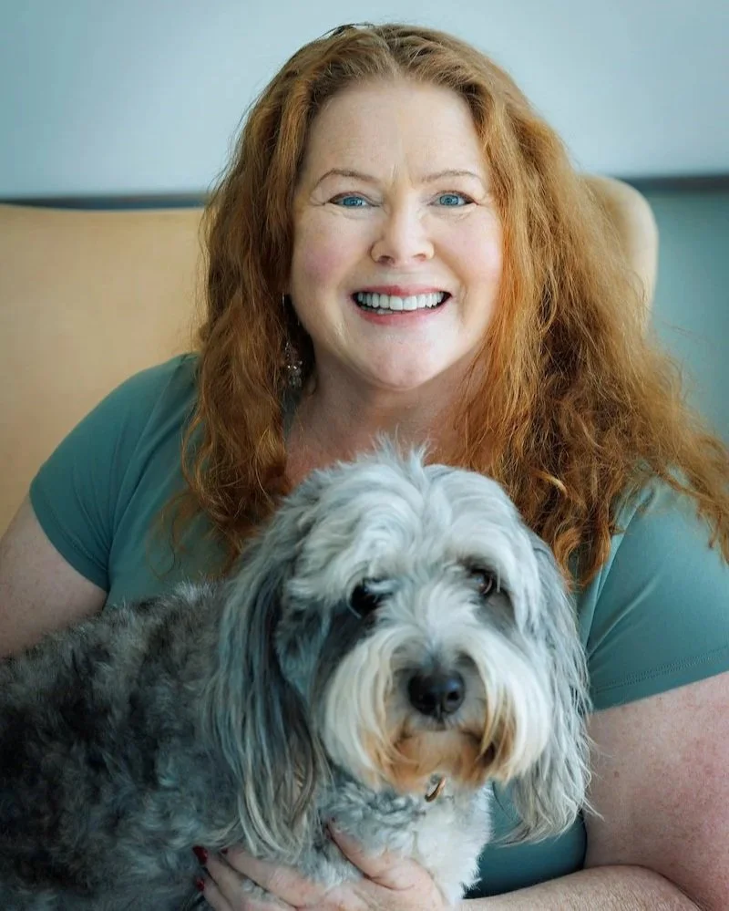 A woman with red curly hair smiling in front of a plain light-colored background.