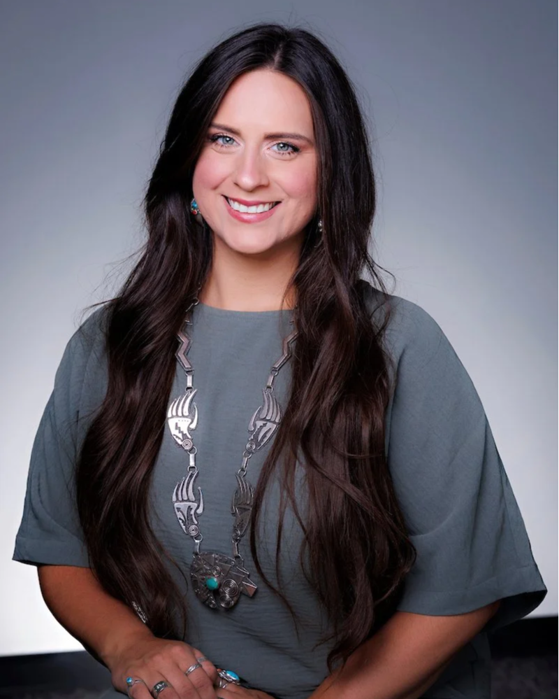 A woman with long brown hair, blue eyes, and wearing makeup, including lipstick, is sitting on a wooden chair with a light neutral-colored backrest. She is smiling subtly with her chin resting on her right hand and appears to be in a bright indoor space with light blue walls.
