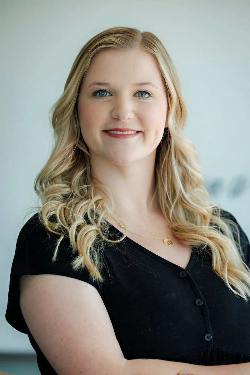 A portrait of a smiling woman with long blond curly hair, wearing a black top and a gold necklace, standing against a neutral background.