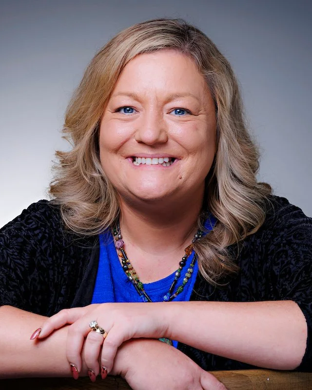 A smiling woman with shoulder-length light brown hair, wearing a black blazer over a blue top, sitting in a tan armchair with crossed hands, in a professional setting.